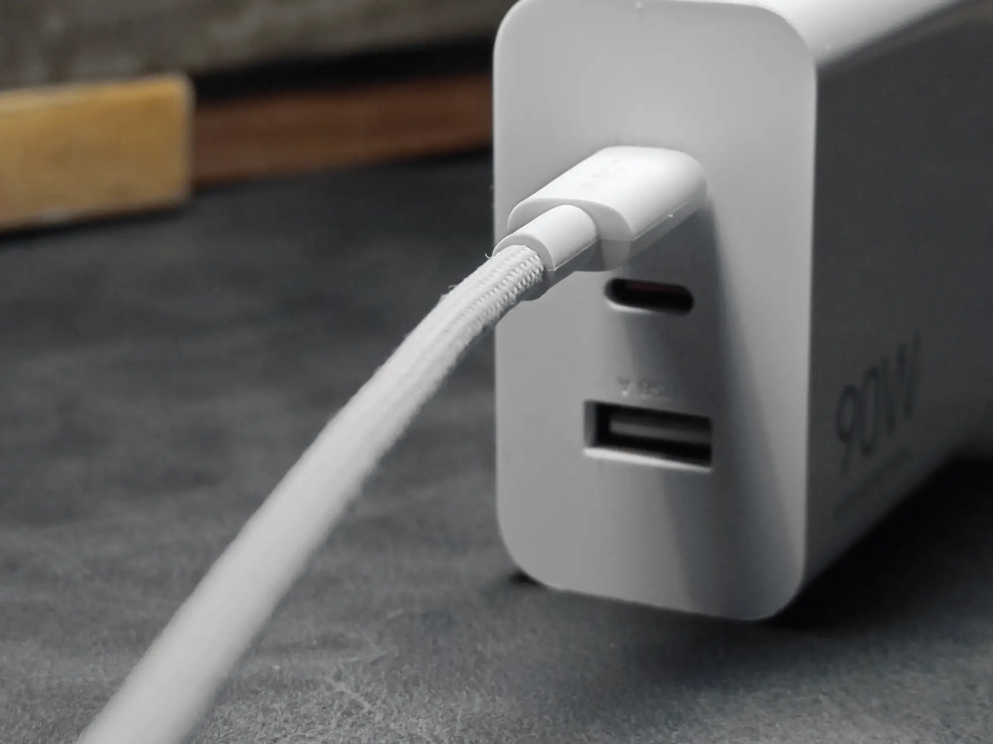 白い90W充電アダプターのUSB-Cポートに、白い布巻きのUSBケーブルが接続されているクローズアップ写真。アダプターの側面には「90W」の文字とUSB-Aポートが見える。充電器の使用例を示す画像。