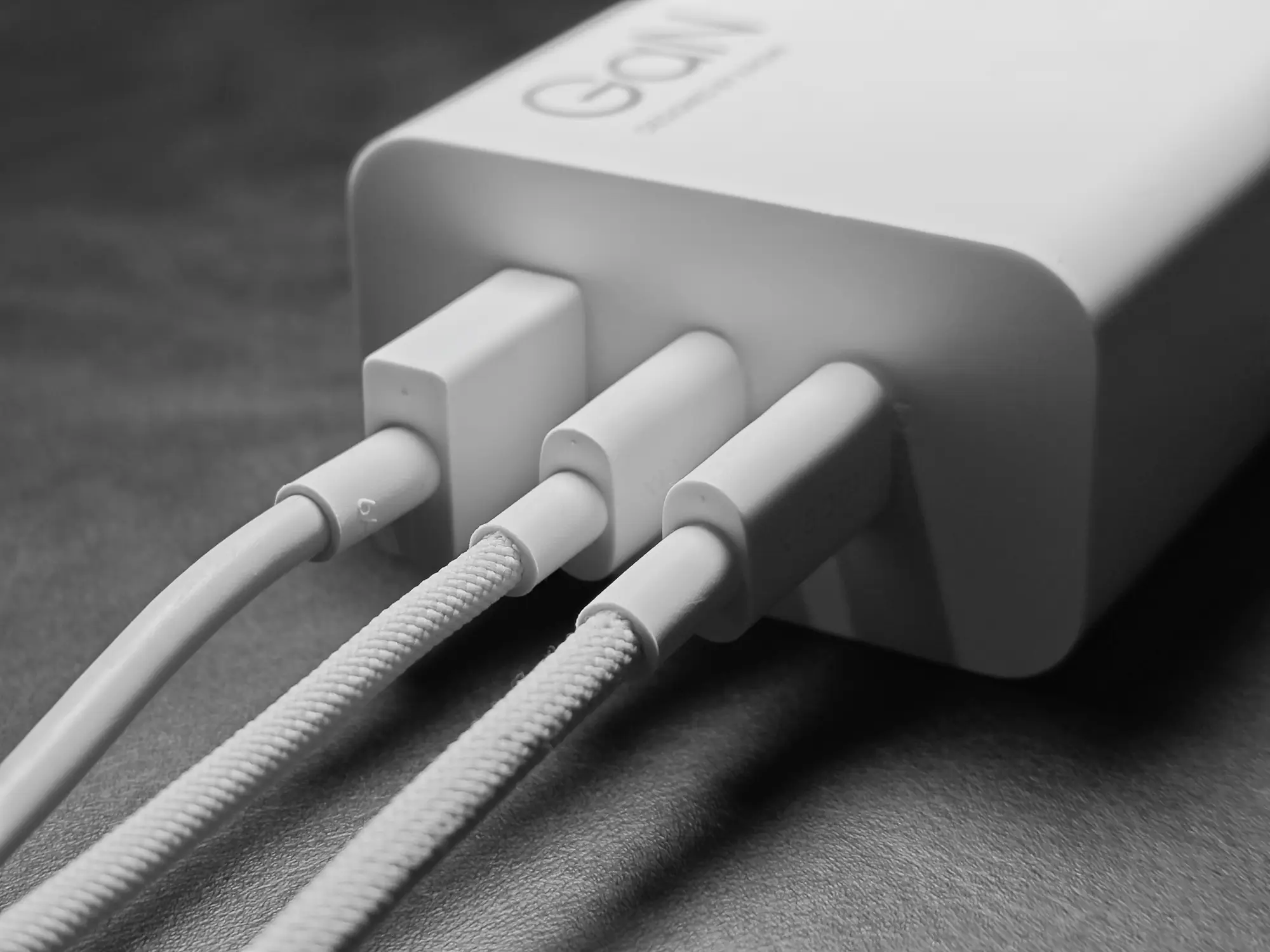 モノクロ写真。白いGaN充電アダプターに、USB-A、USB-Cの全3ポートすべてにケーブルが接続されているクローズアップ。アダプター本体には「GaN」の文字が見える。3台同時充電の使用例を示す画像。