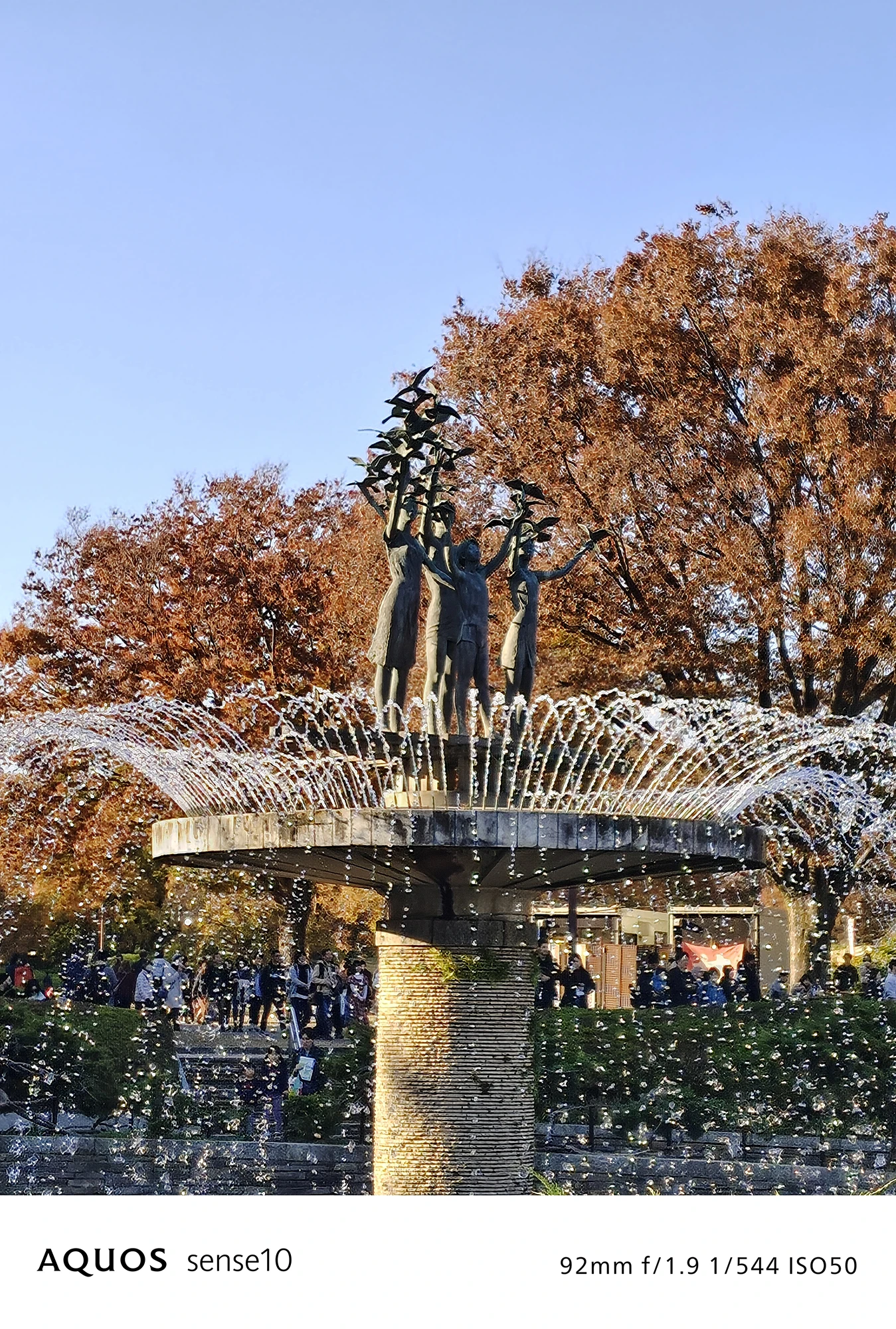 AQUOS sense10撮影サンプル：2倍ズームで捉えた**紅葉に囲まれた噴水と像**（f/1.9 ISO50）