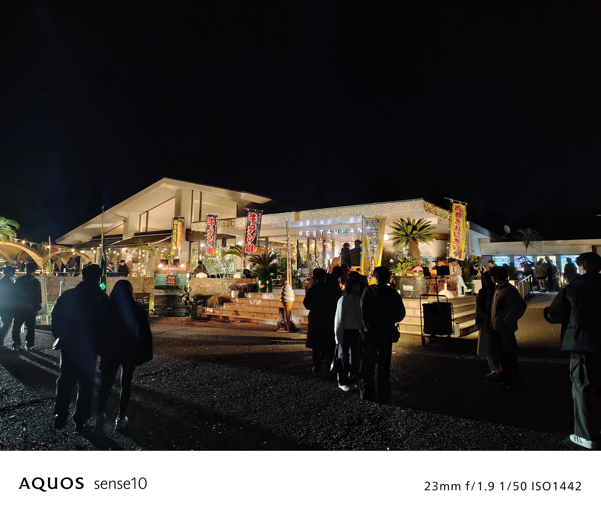 AQUOS sense10撮影サンプル：広角で捉えた**夜のレストラン（休憩所）と集まる人々**（f/1.9 ISO1442）