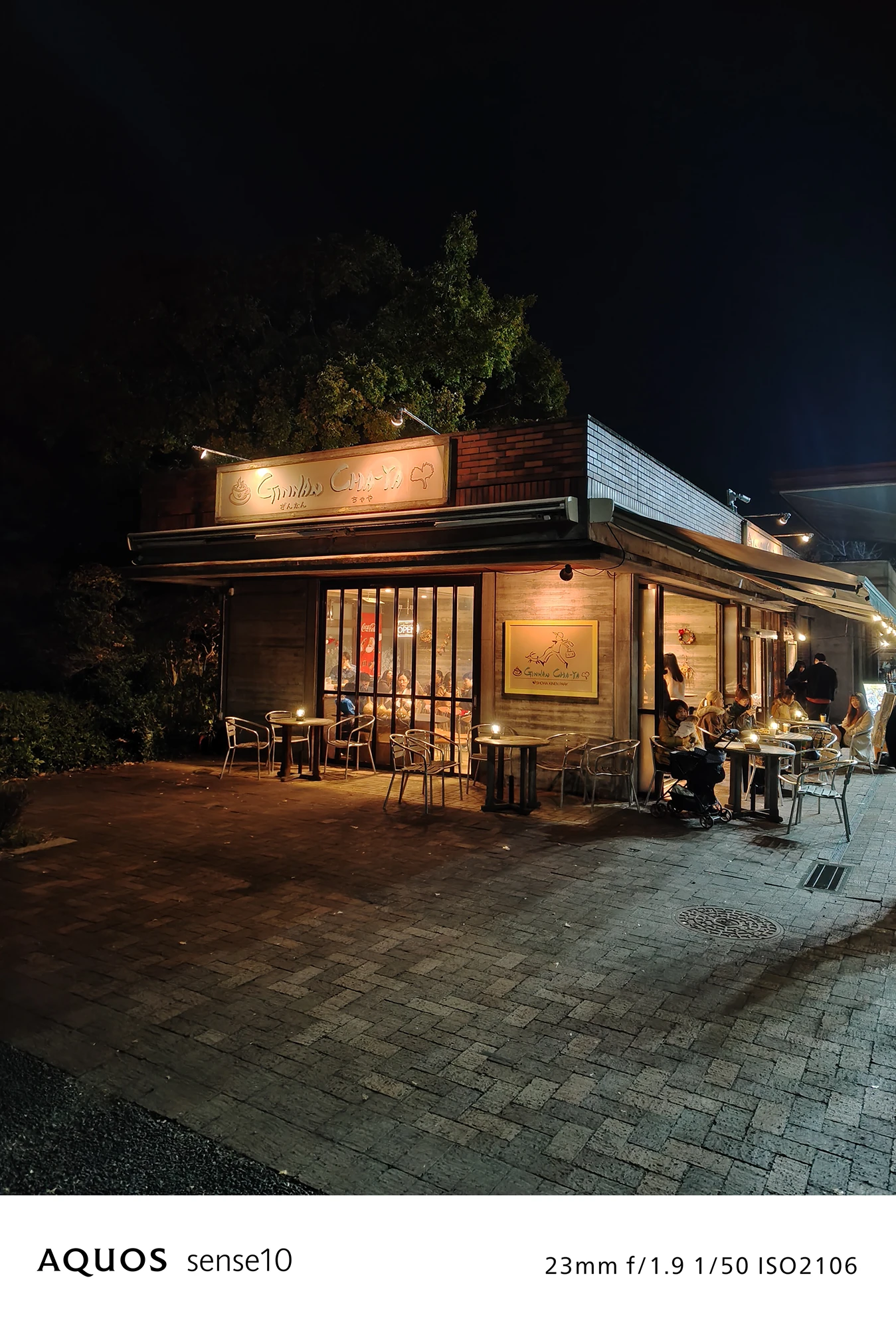 AQUOS sense10撮影サンプル：**レンガ調の建物とテラス席**がある、夜の公園の休憩スポット（f/1.9 ISO2106）