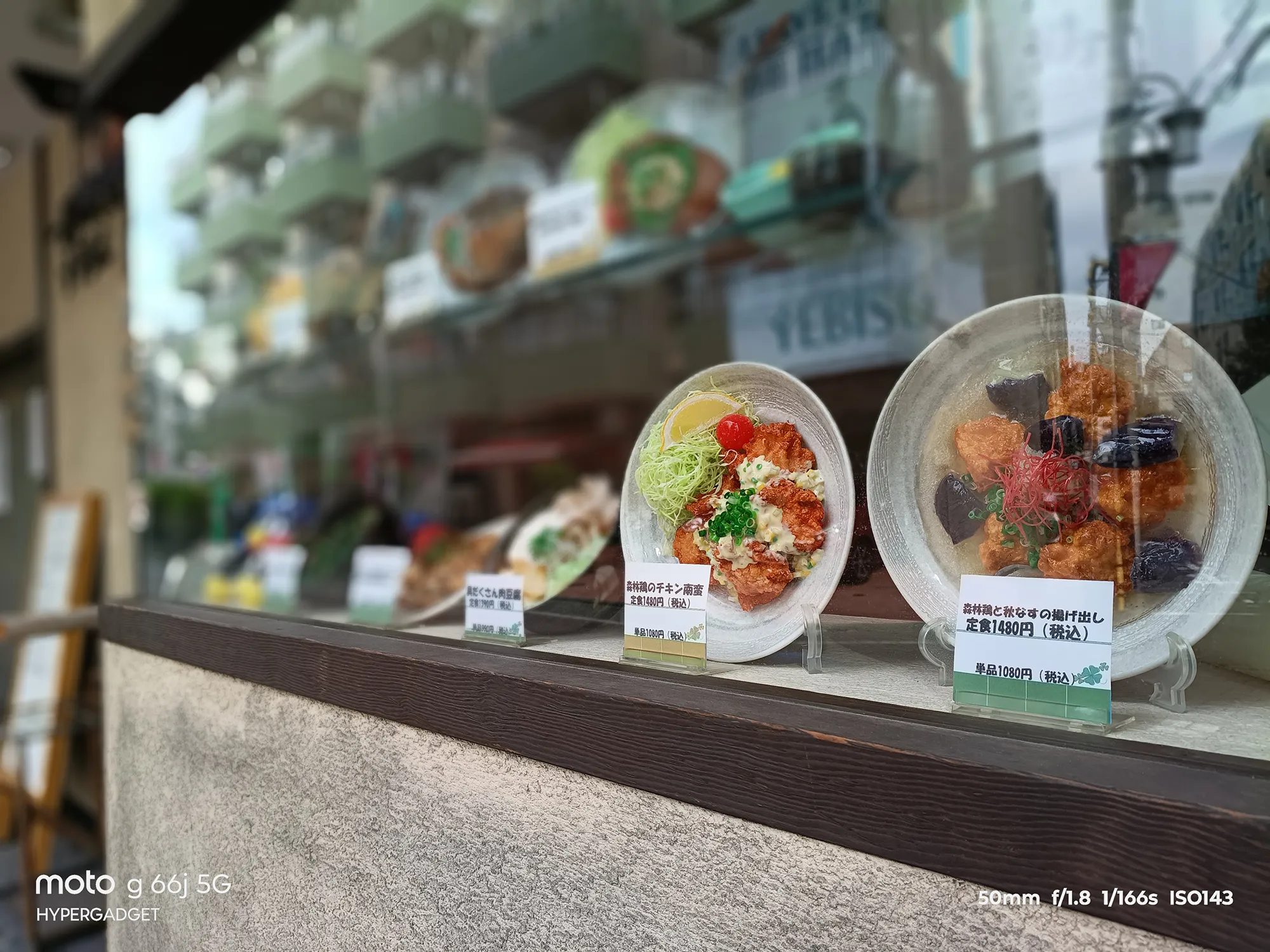 moto g66j 5Gのカメラ作例：食品サンプル（チキン南蛮定食など）をガラス越しに撮影。背景のボケ味と料理の質感をレビュー