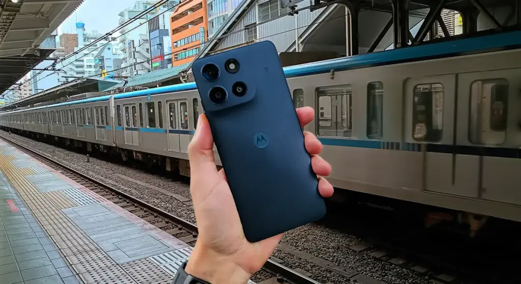 moto g66j 5Gの本体背面とカメラモジュールを手に持った写真。走行中のJR電車が背景に写り、カメラ性能レビュー用であることを示す