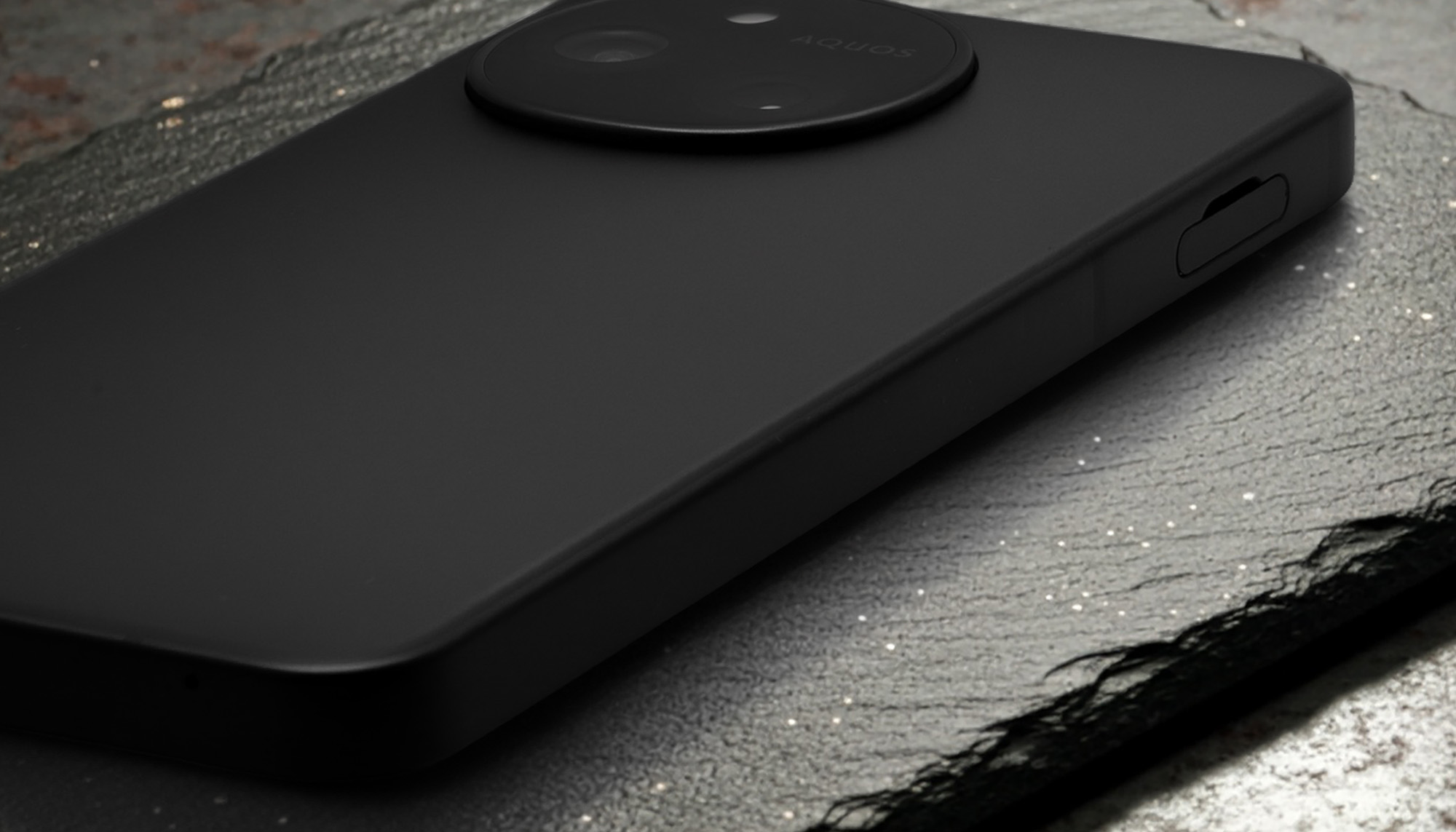 AQUOS sense10またはSH-M33のマットブラックの背面を接写したクローズアップ。本体表面の均一で滑らかな質感が強調され、上部には円形のカメラモジュールの縁、右側面には電源ボタンが見える。プレミアム感のあるデザインと、高い塗装品質をアピール。
