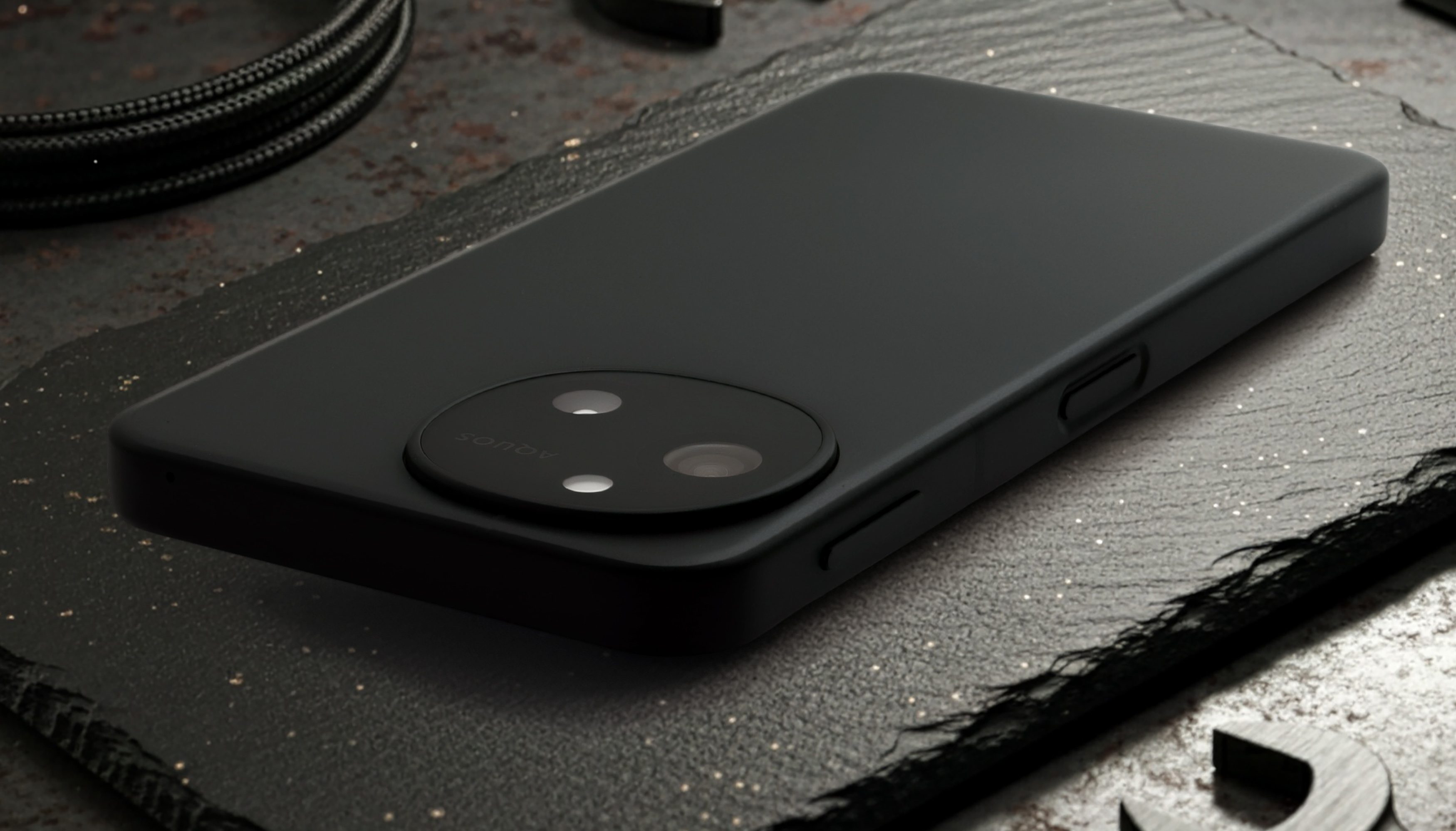 AQUOS sense10の背面製品写真。本体はマットブラックで、左上には円形のデュアルカメラモジュールとロゴが見える。側面には音量ボタンと電源ボタンが配置されており、落ち着いたデザインと高い質感を強調する。背景には工具やケーブルが配置されている。