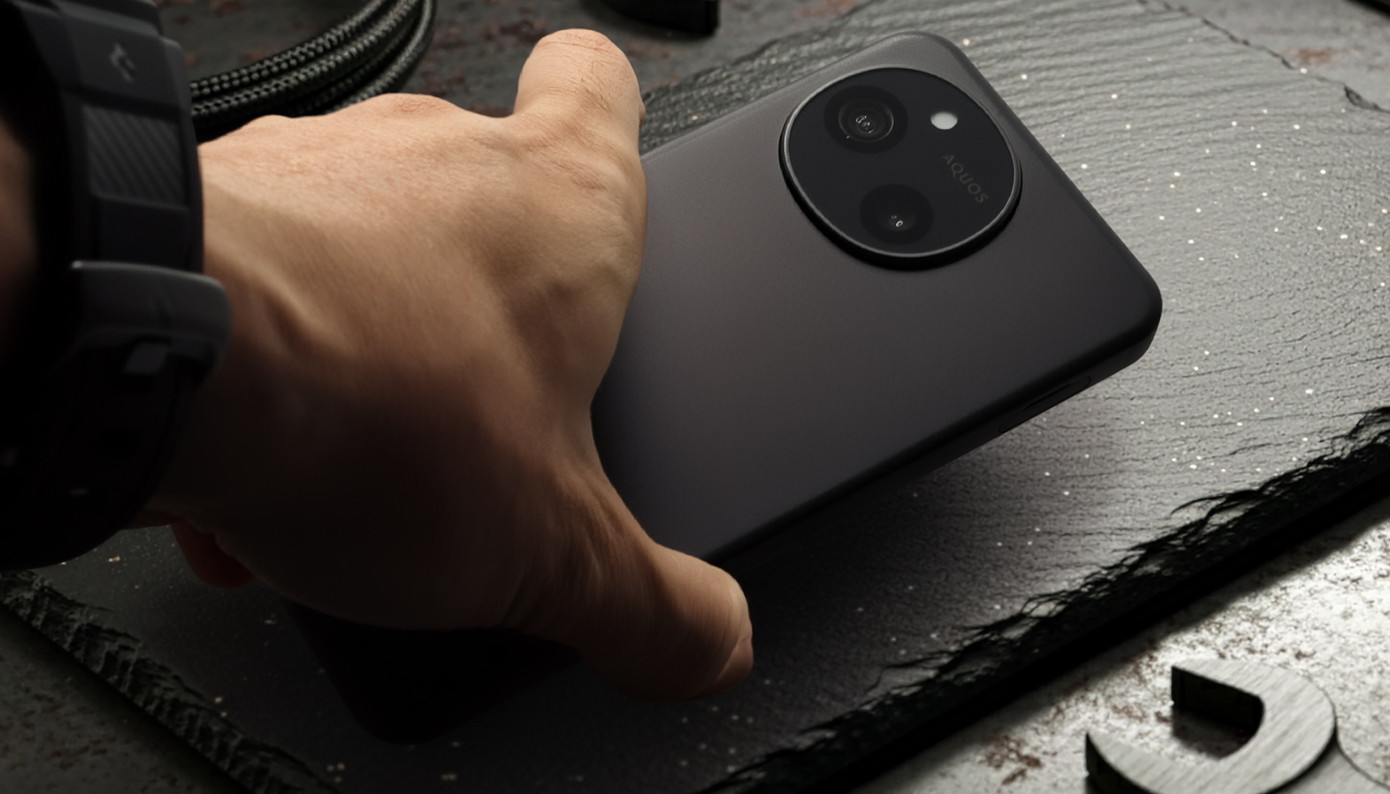 AQUOS sense10またはSH-M33のマットブラックの背面を、手が握っているクローズアップ写真。特徴的な円形のデュアルカメラモジュールと「AQUOS」ロゴがはっきりと見える。本体のサイズ感と、マットな質感による優れたグリップ性や高いホールド感をアピール。背景には工具が見える。