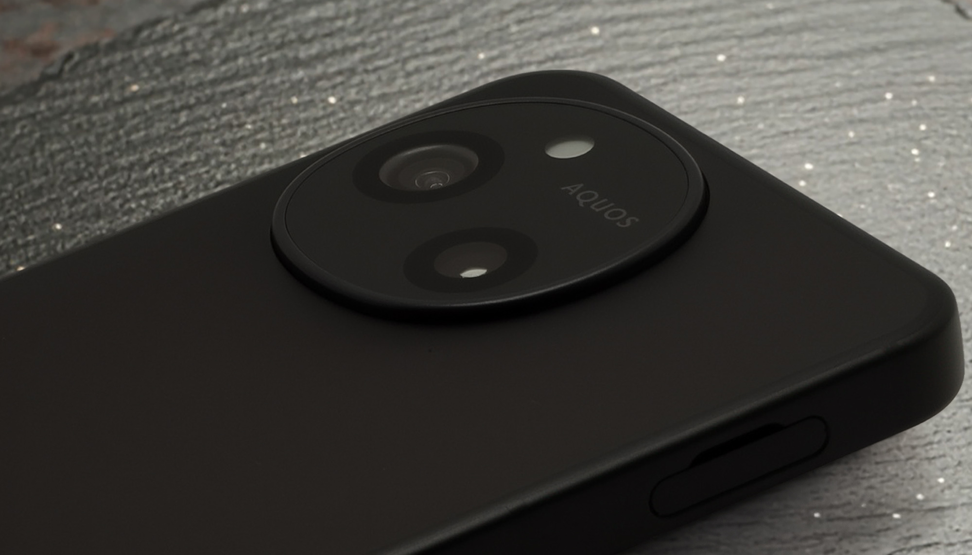 AQUOS sense10（またはSH-M33）の背面、円形カメラモジュールを接写した写真。カメラユニット内にはデュアルカメラとフラッシュ、そして「AQUOS」のロゴが見える。本体はマットブラックで、側面の電源ボタンがわずかに確認できる。特徴的なカメラデザインと高い質感をアピール。