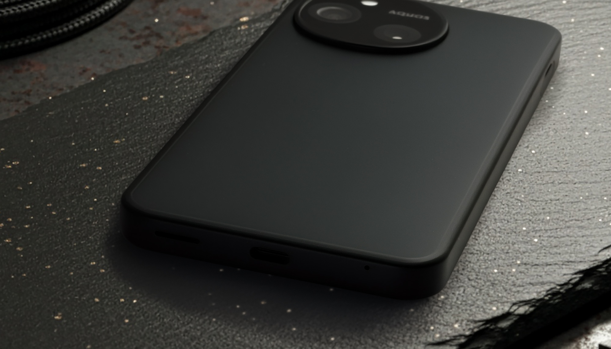 AQUOS sense10またはSH-M33のマットブラックの背面と底面を接写した写真。底面にはUSB Type-Cポートとマイクまたはスピーカーグリルが見える。上部には円形のカメラモジュールと「AQUOS」ロゴが配置されており、本体の質感と端子の配置を示す。