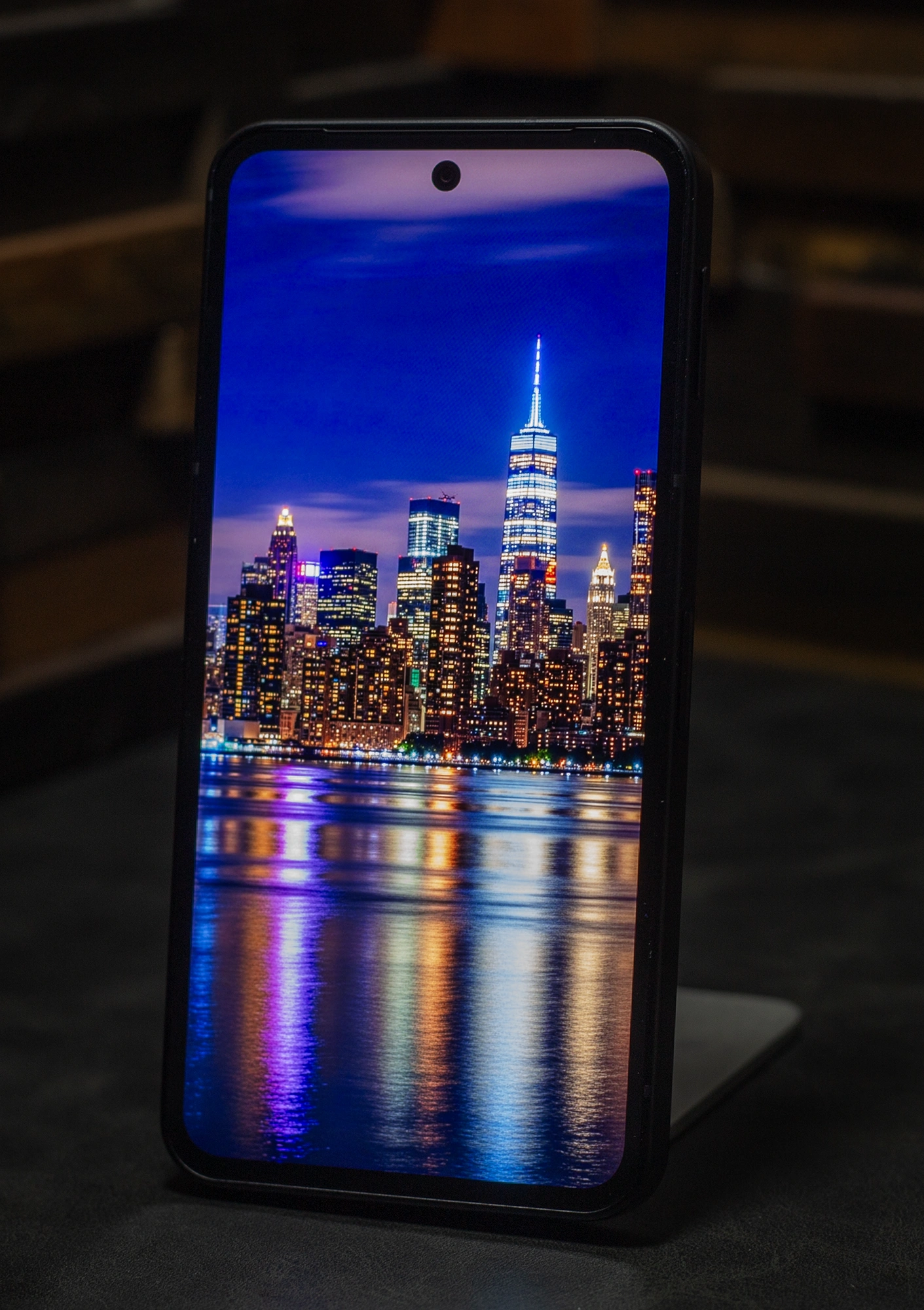 スマートフォン画面に表示された、ニューヨークやシカゴのような水辺の高層ビル夜景。水面に映る光の反射が鮮明で、有機ELディスプレイの深い黒と鮮やかな発色を強調。特に夜間表示における高いコントラストと没入感を示すサンプル。