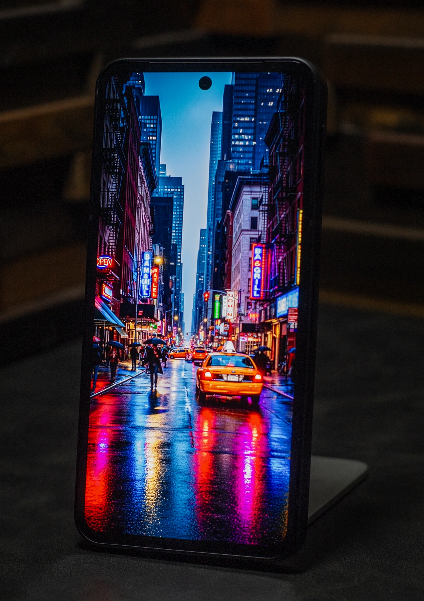 スマートフォン本体に表示された、雨に濡れた路面にネオンとイエローキャブが反射するニューヨークの夜景。高輝度で彩度の高い有機ELディスプレイの特性を強調しており、特に光の映り込みとコントラスト表現の優秀さを示す。狭いベゼルと画面上部のパンチホールカメラが見える。