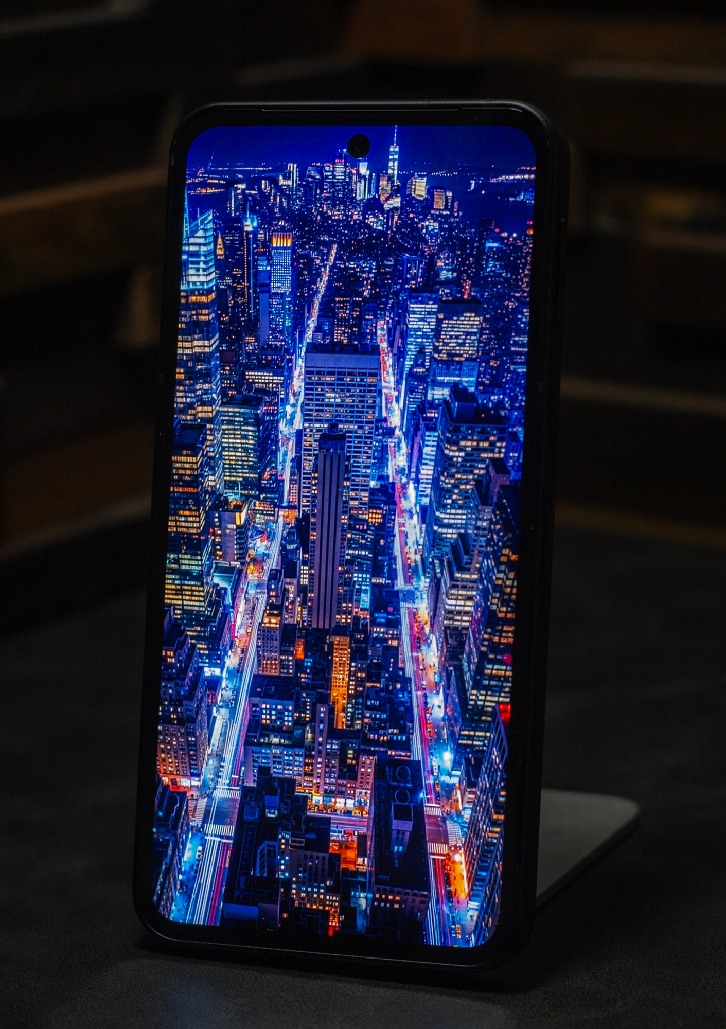スマートフォン本体に表示された、ニューヨークや東京のような高層ビル群の空撮夜景。青とオレンジの光のコントラストが際立ち、有機ELディスプレイの黒の沈み込みと色彩表現の深さを強調。縦長の画面いっぱいに表示された没入感のあるデザインで、パンチホールカメラが見える。