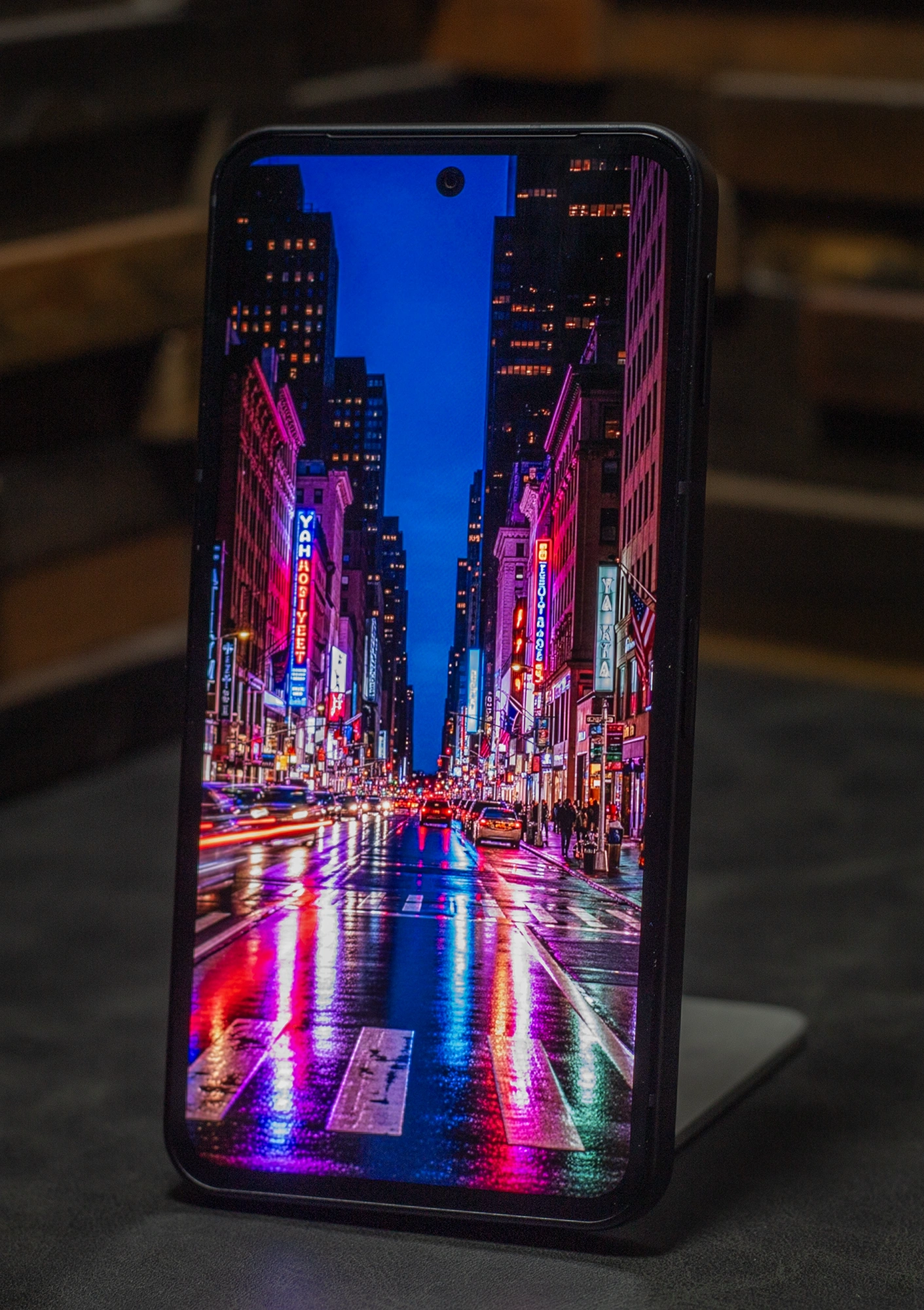 スマートフォン本体に表示された、雨上がりのニューヨーク・タイムズスクエアのようなネオン輝くストリート夜景。濡れた路面への光の反射が鮮烈で、高輝度で彩度の高い有機ELディスプレイの発色性能を強調。上下左右の狭いベゼルとパンチホールカメラのデザインが見える。