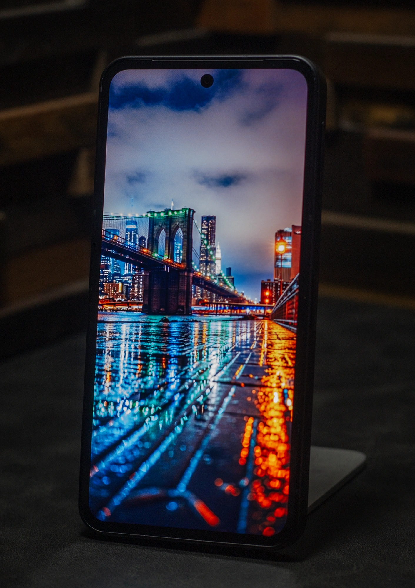 スマートフォン本体に表示された、ブルックリン橋とマンハッタンの夜景の壁紙。水面にネオンが反射し、画面の高いコントラストと鮮やかな色再現性を強調している。パンチホール式のフロントカメラが見える狭ベゼルのデザインで、有機ELディスプレイの視認性を示す。