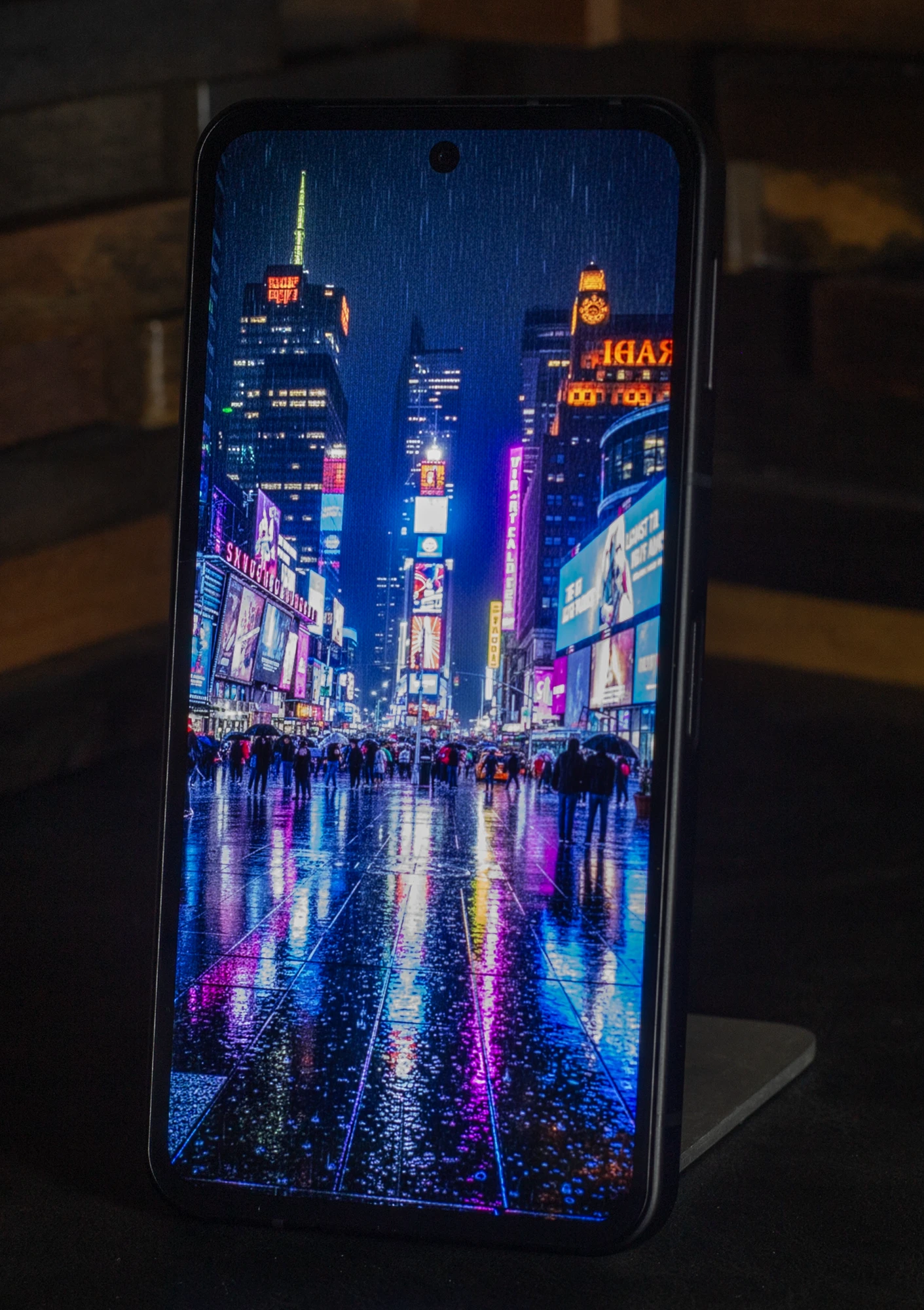 スマートフォン本体に表示された、雨の日のタイムズスクエアの夜景。ネオンサインと路面の水たまりへの鮮烈な光の反射が描かれており、有機ELディスプレイの広い色域と高輝度表示性能を強調。上下左右のベゼルが狭く、高い没入感とパンチホールカメラのデザインが見える。