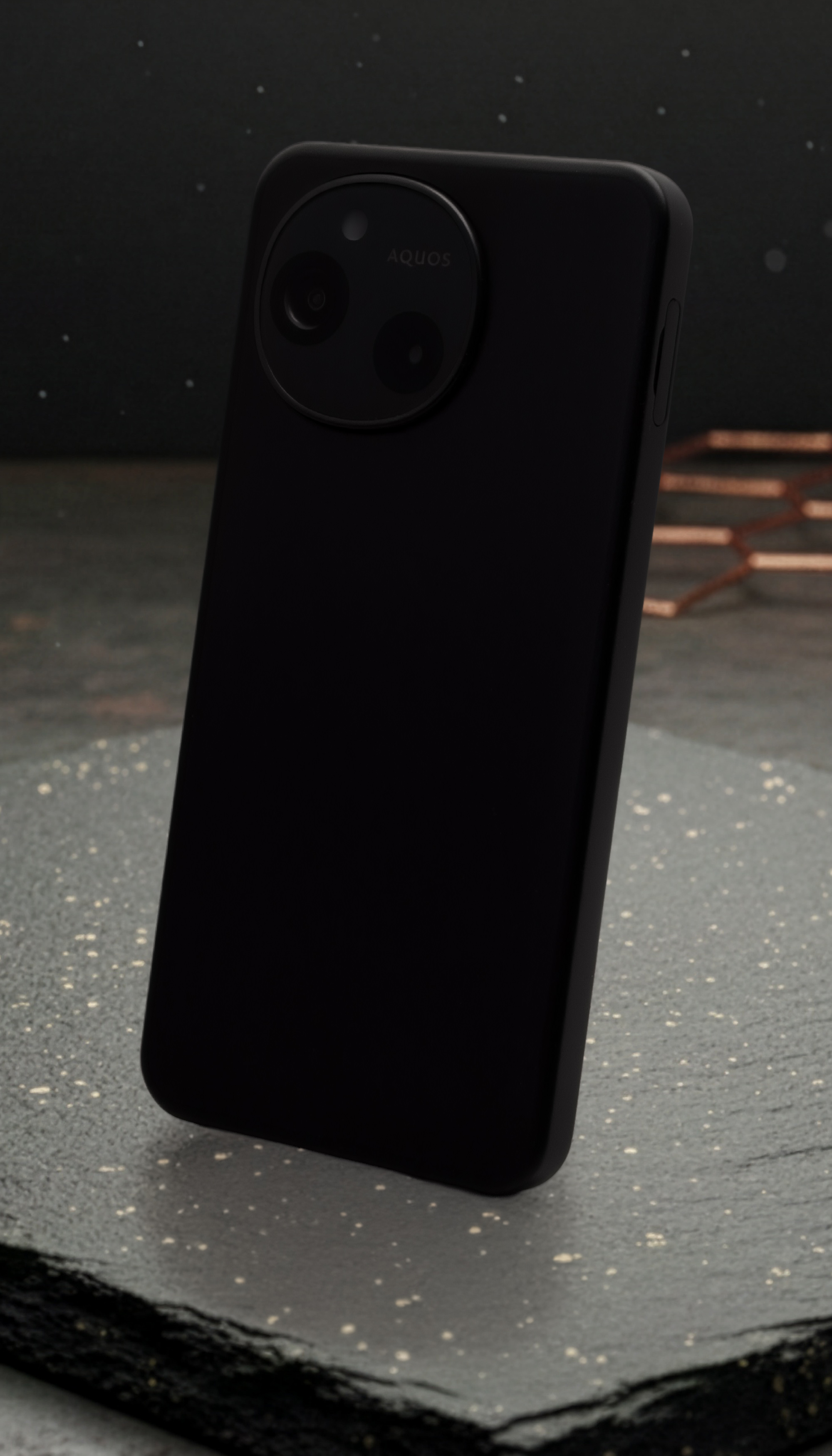 AQUOS sense10またはSH-M33の背面デザインを示す製品写真。マットブラックの本体は石のような質感の台座に立てかけられ、上部には円形のデュアルカメラモジュールと「AQUOS」ロゴが配置されている。シンプルで洗練されたデザインと、カメラユニットの独自性を強調。