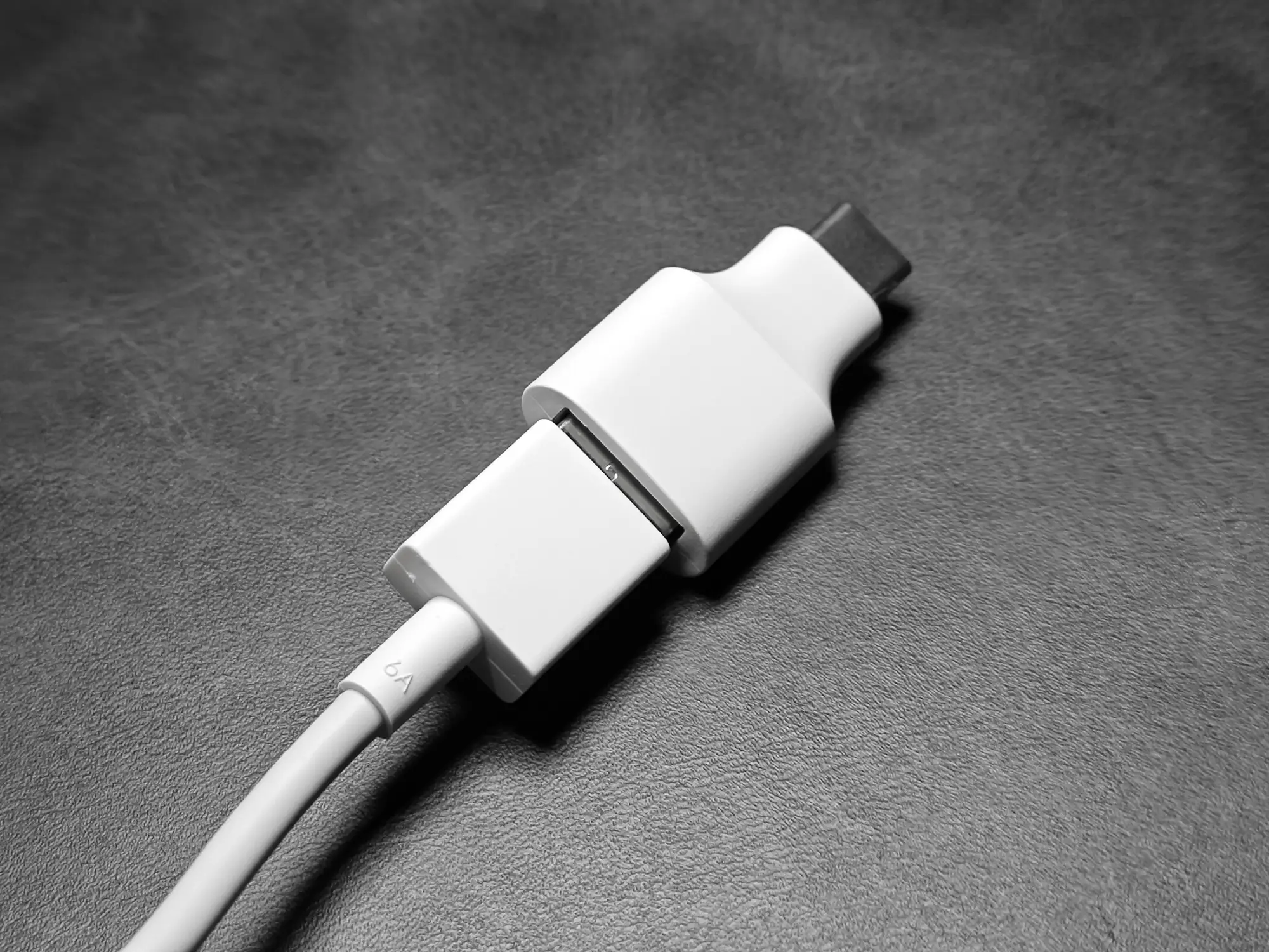 モノクロ写真。白いUSBケーブルに、四角い白いアダプター（USBテスターまたは延長パーツ）が接続され、さらにその先に別のコネクタが挿入されている。ケーブルには「6A」の文字が見える。USB充電のテストや計測に使用される接続状態。