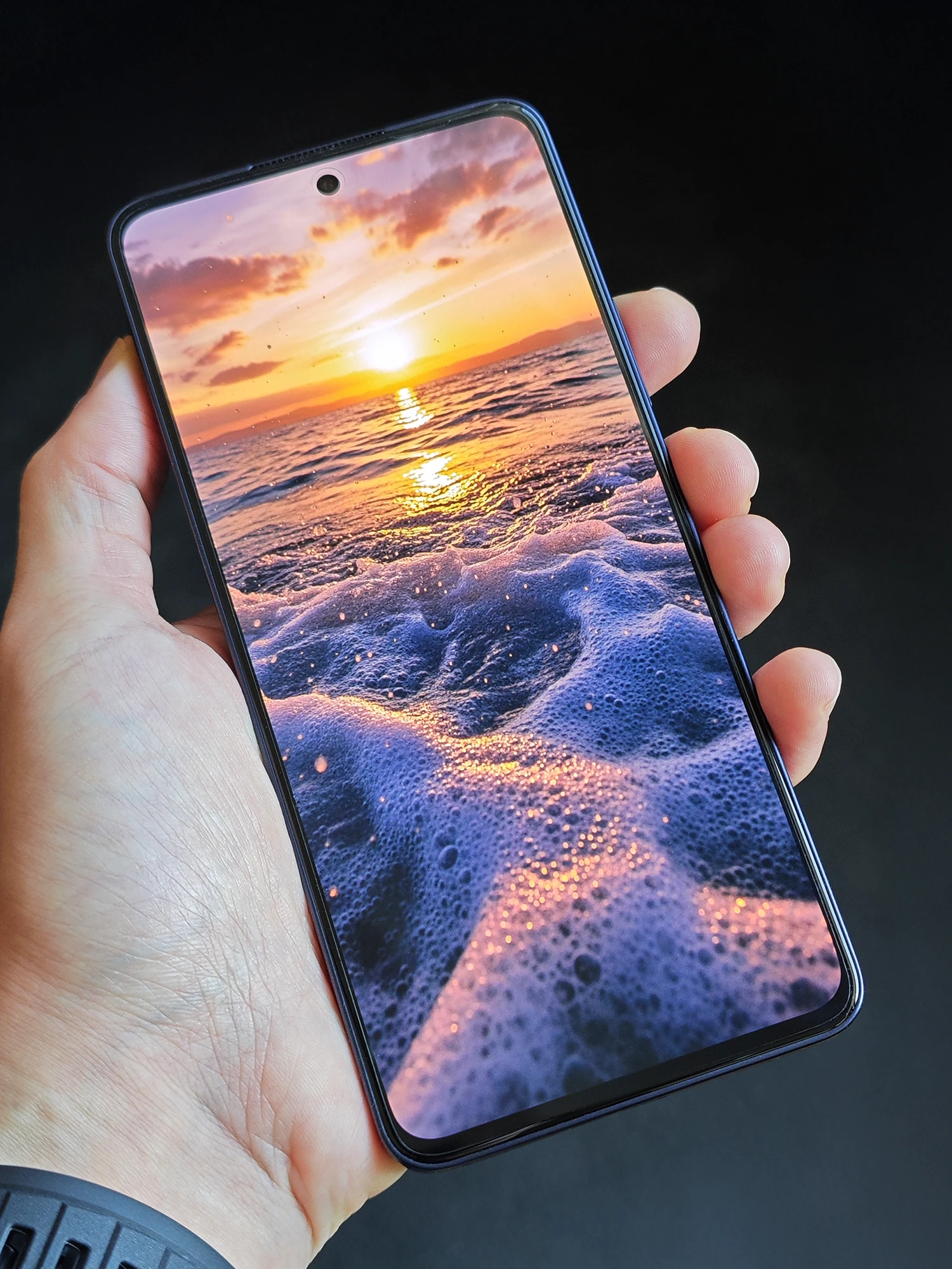 OPPO Reno13 Aを手に持ち、ディスプレイに鮮やかな夕日の波打ち際を表示した様子