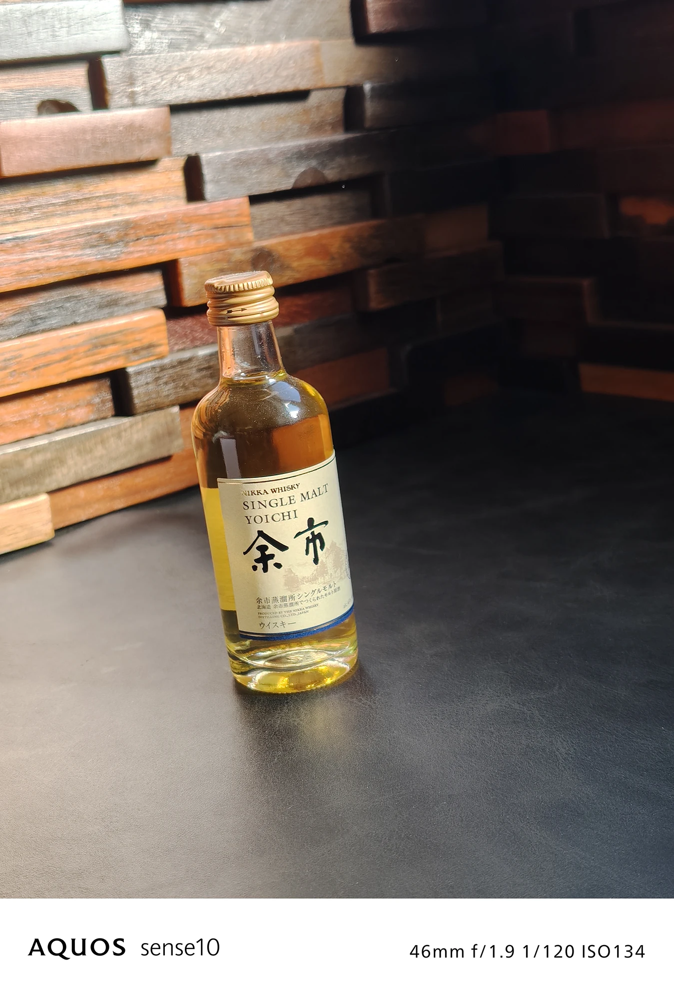 AQUOS sense10で撮影された、「余市（YOICHI）」シングルモルトウイスキーのミニボトルのクローズアップ写真。46mm f/1.9 1/120 ISO134の撮影情報が付記されている。ボトルのラベルの文字や液体の透明感が鮮明に表現され、被写体認識と質感表現に優れたカメラ性能を示す。背景の木目壁と黒いテーブルの質感も捉えている。