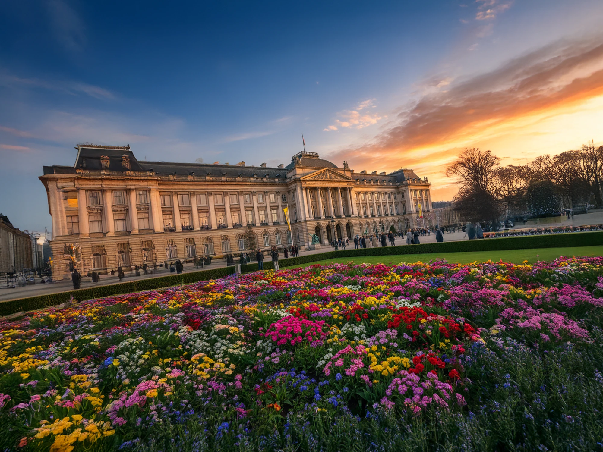 色とりどりの花が広がる庭園越しに宮殿を望む夕景。建物の重厚な外観と鮮やかな花畑、温かい夕焼け空が調和した風景。