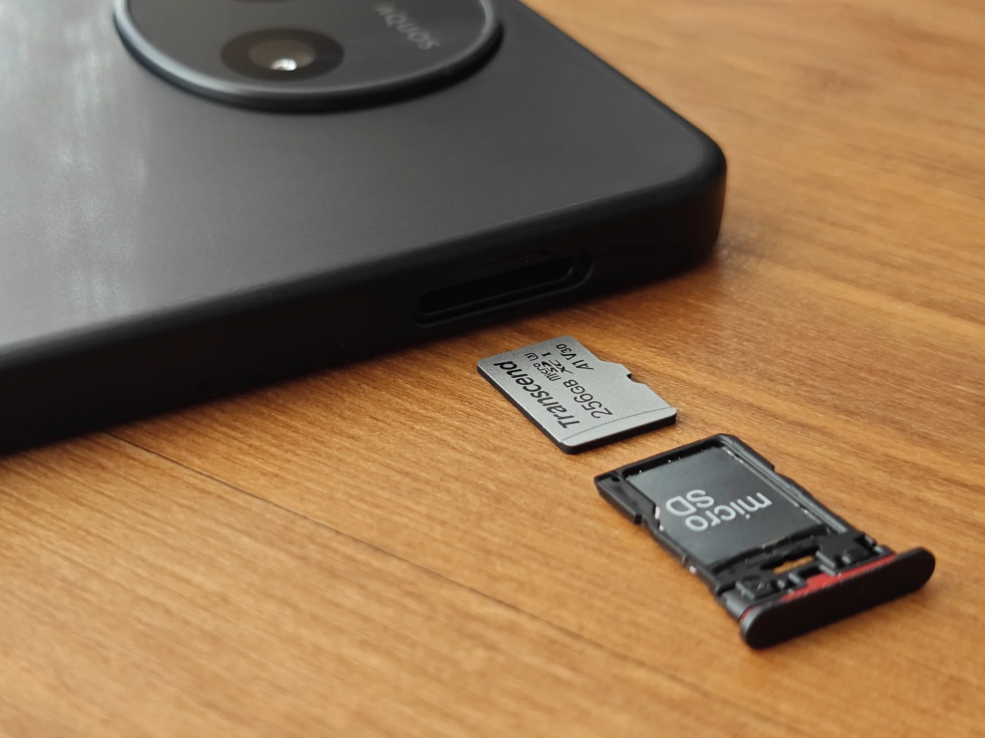 AQUOS sense10または類似スマートフォンの底面付近を接写した写真。本体横から取り出されたMicro SDカードトレイと、Transcend製256GBのMicro SDカードが見える。外部ストレージ（Micro SD）による容量拡張に対応しており、データ保存や写真・動画の大量保存に優れていることをアピール。