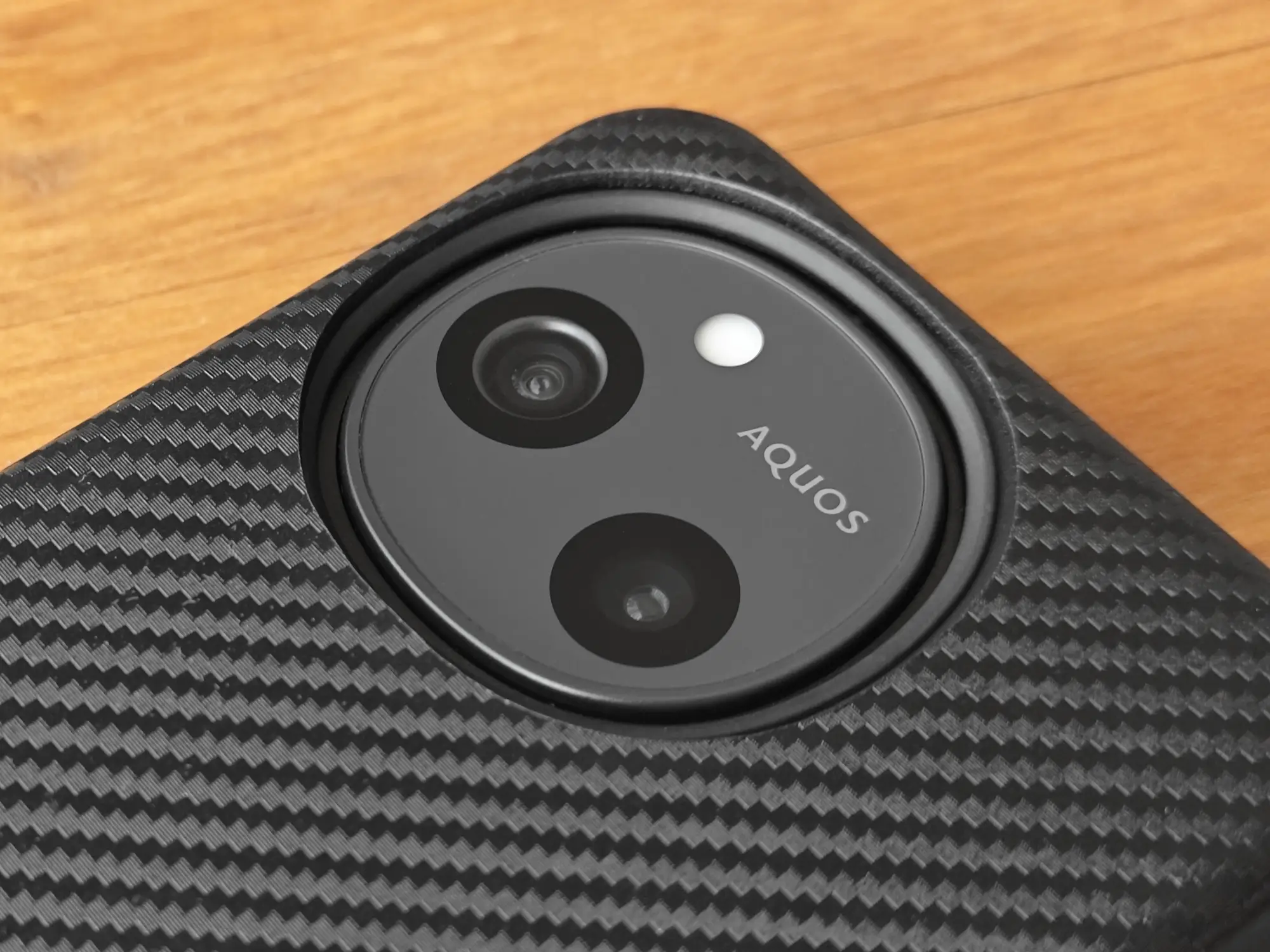 カーボン調のスマートフォンケースを装着したAQUOS sense10またはSH-M33の背面を接写。ケースはカメラモジュールを円形にくり抜いて保護しており、内部のAQUOSロゴ入りデュアルカメラがはっきりと見える。カメラユニットの保護性能と、カーボンケースの高いデザイン性をアピール。