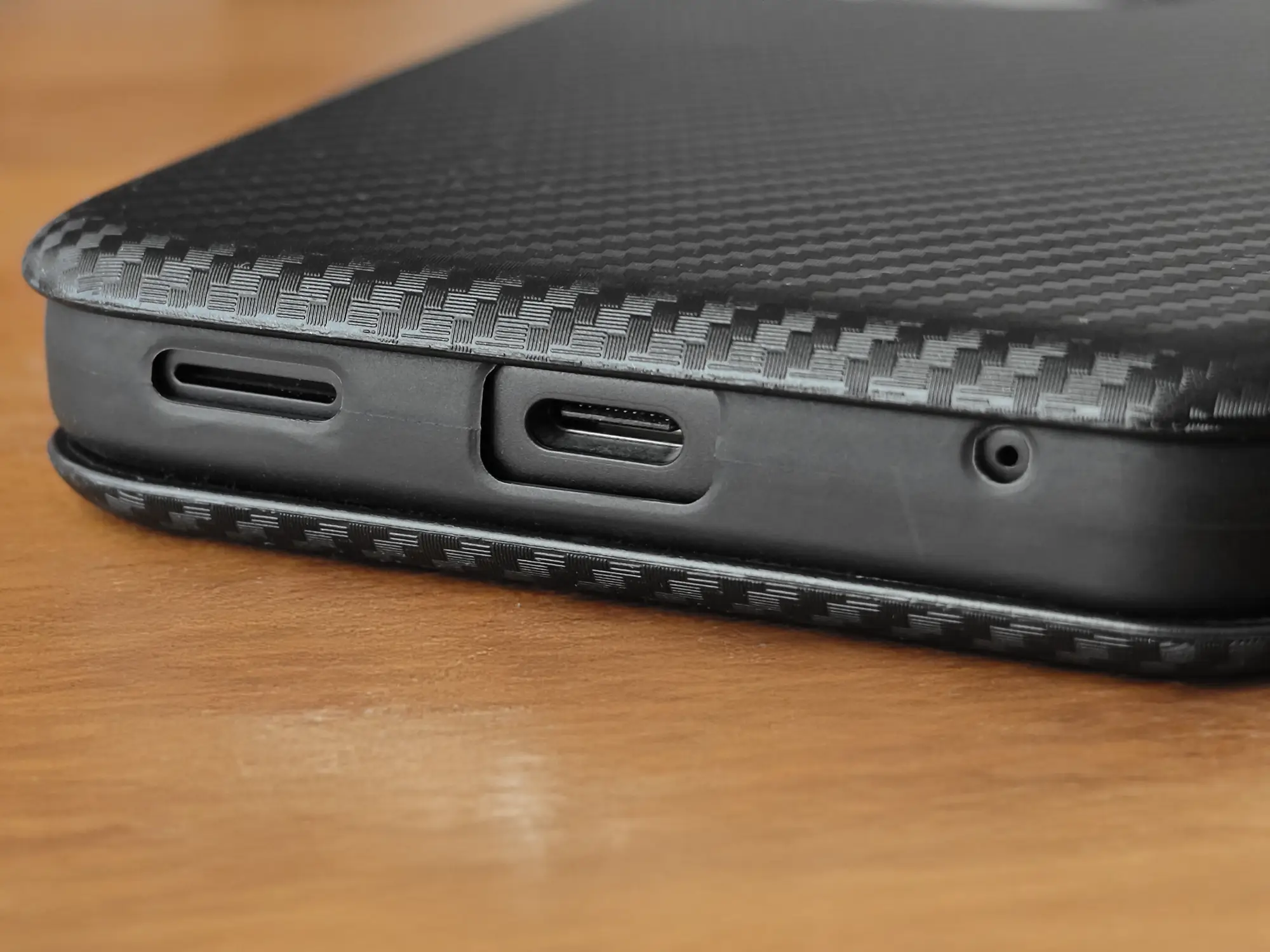 カーボンファイバー調のケースを装着したスマートフォンの底面を接写。ケースは、USB Type-Cポート、スピーカーグリル、マイク穴の各開口部を正確にくり抜いており、ケース装着時でも機能へのアクセスが容易であることを示す。高い保護性能と機能性を両立したスマートフォンケースのサンプル。