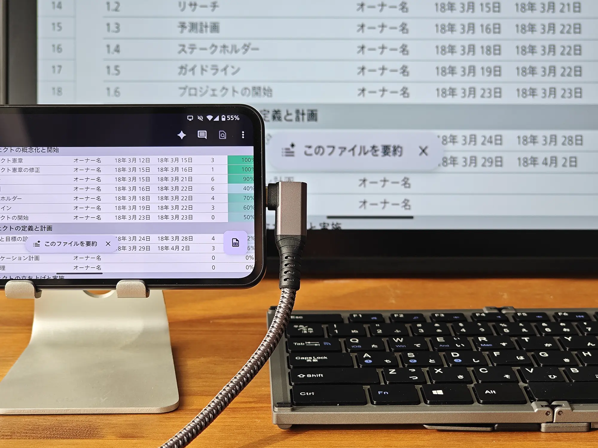スマートフォンがUSB-Cケーブルを介して外部ディスプレイに接続され、画面出力（ミラーリングまたはデスクトップモード）を行っている様子。スマートフォンと外部ディスプレイには、プロジェクト管理の表のようなExcel/スプレッドシートの画面が表示されている。有線での外部ディスプレイ出力機能と、モバイルデバイスでのマルチタスク、ビジネス用途への適性を示す。