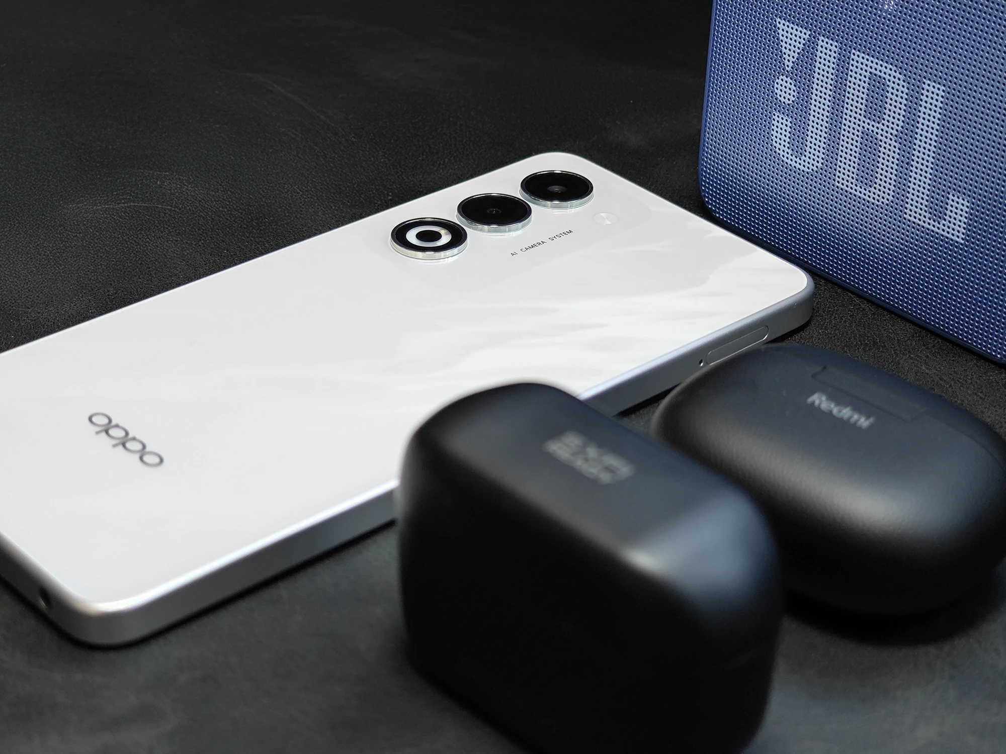 ホワイトの背面を持つOPPO A5 5Gと、右奥にJBLの青いポータブルスピーカー、手前にRedmiワイヤレスイヤホンの充電ケースが並べられた、モバイルオーディオセットのクローズアップ写真。