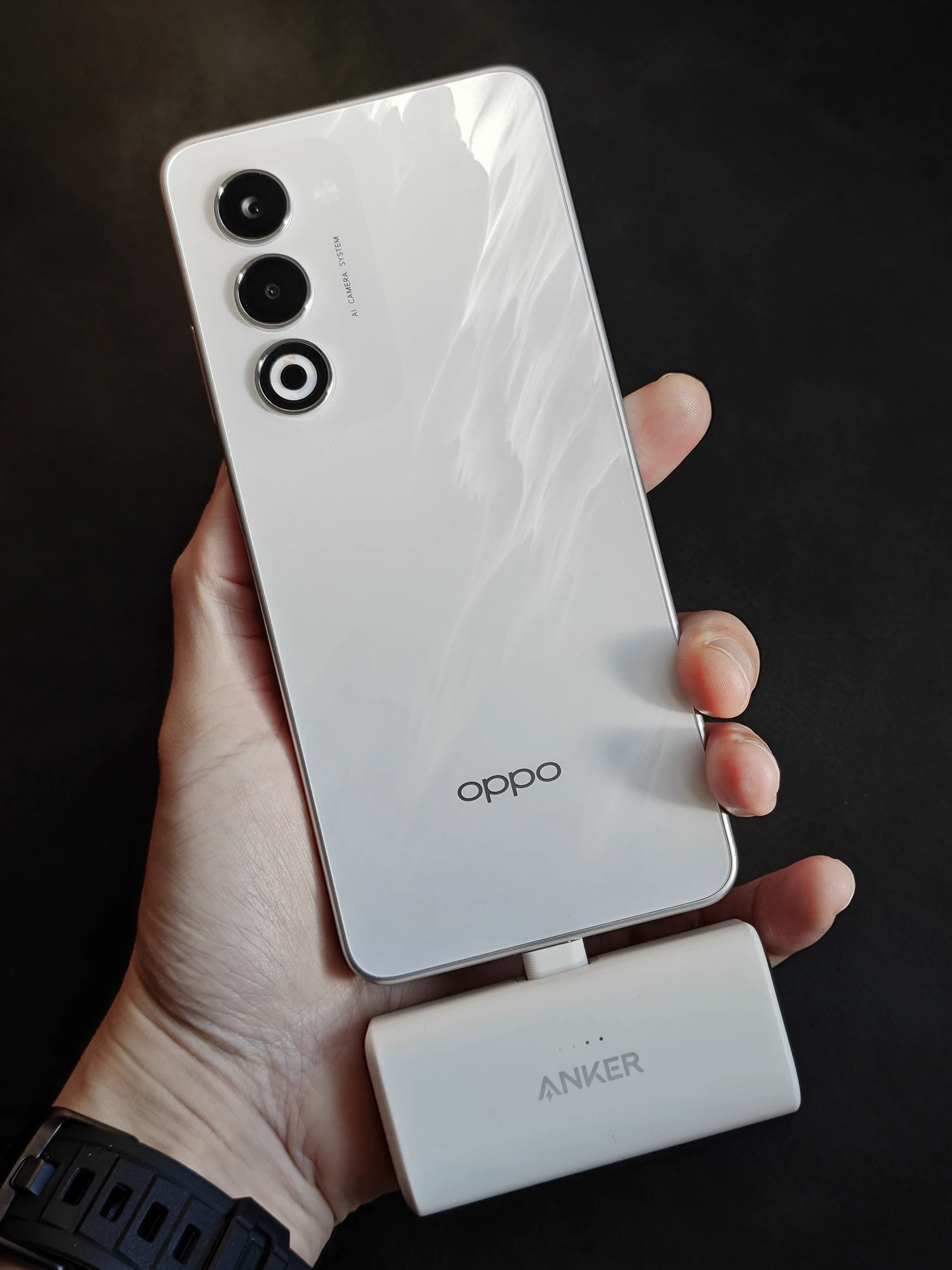 白いOPPO A5 5Gの背面を手で持っている様子。底部にはAnker製の白い小型モバイルバッテリーが接続されている。光沢のある特殊加工の背面と、縦に並んだトリプルカメラが特徴。モバイルバッテリーを使った充電の容易さと、スタイリッシュなデザインをアピール。