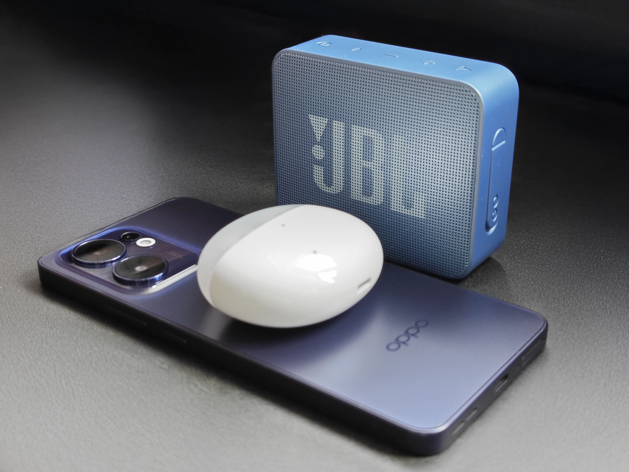 OPPO Reno13 Aの上にワイヤレスイヤホンケースを置き、背後にJBLのポータブルBluetoothスピーカーを並べた外観画像
