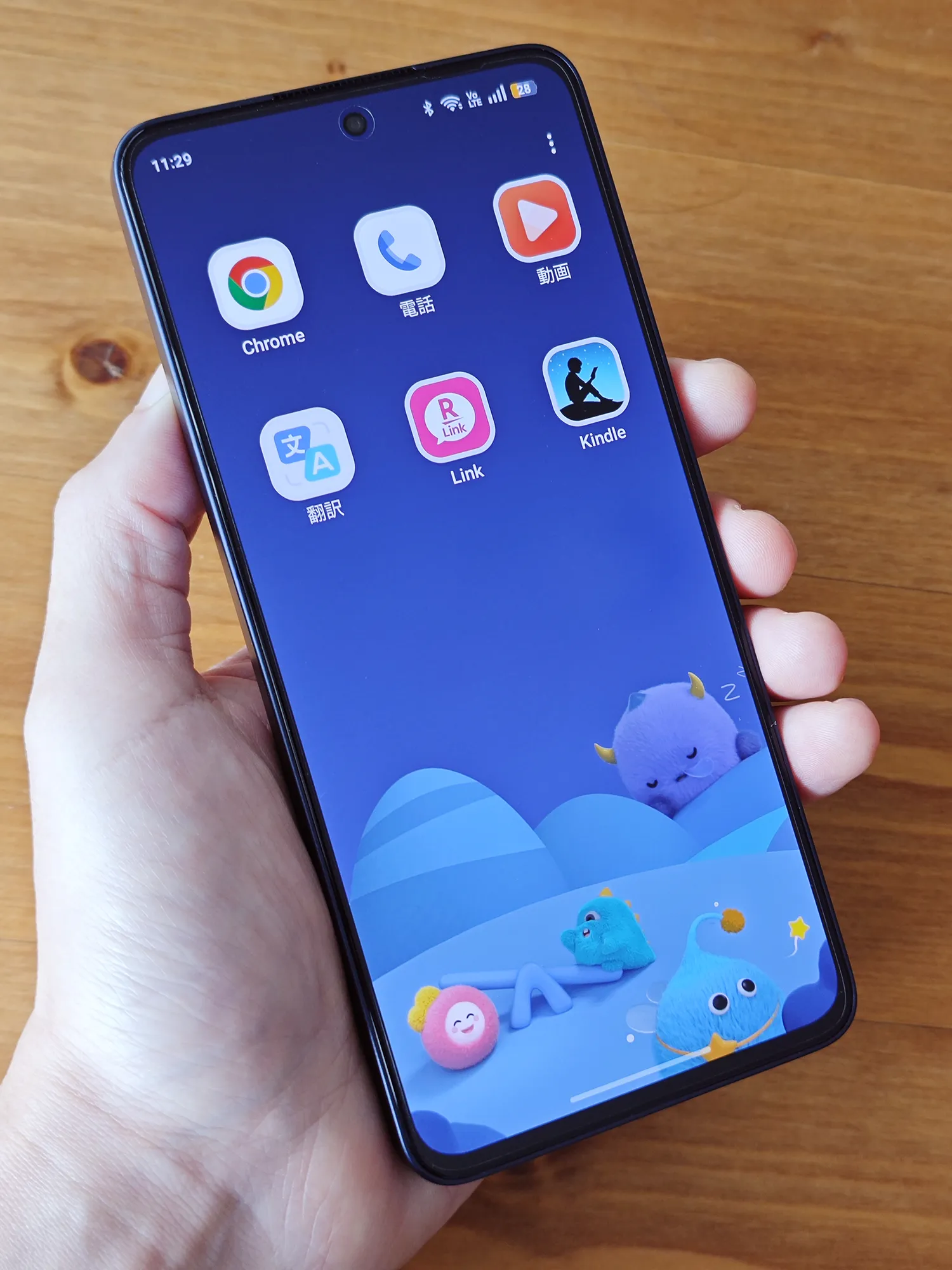 OPPO Reno13 Aを手に持ち、キャラクターが描かれた壁紙のホーム画面を表示している様子。画面にはChrome、電話、動画、翻訳、Rakuten Link、Kindleの計6つのアプリアイコンが並んでいる。