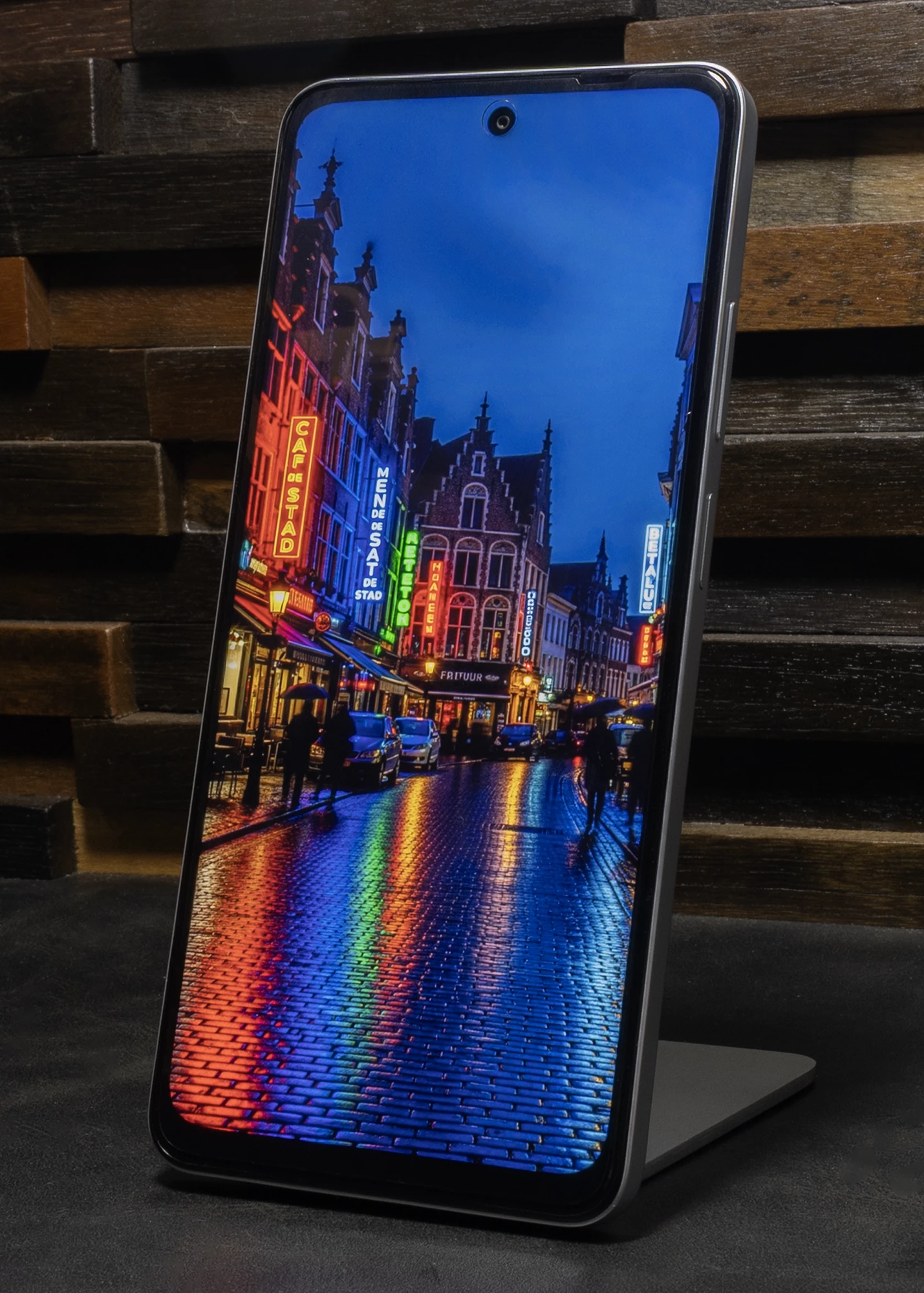 スタンドに立てられたOPPO A5 5Gのディスプレイに、雨の夜のヨーロッパの街並みと、濡れた石畳にネオンサインが反射している写真が表示されている。ディスプレイ上部中央にはパンチホールカメラが見える。