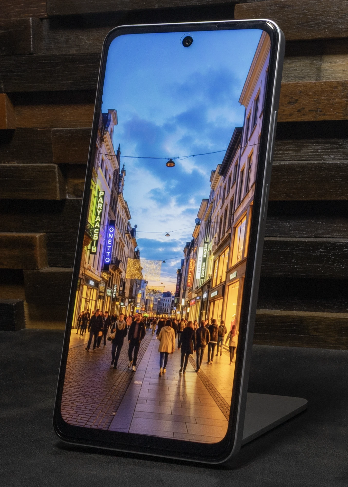 スタンドに立てられたOPPO A5 5Gのディスプレイに、夕暮れ時で歩行者やネオンサインの灯りが賑わうヨーロッパの石畳の通りが表示されている。上部中央にはパンチホールカメラが見える。