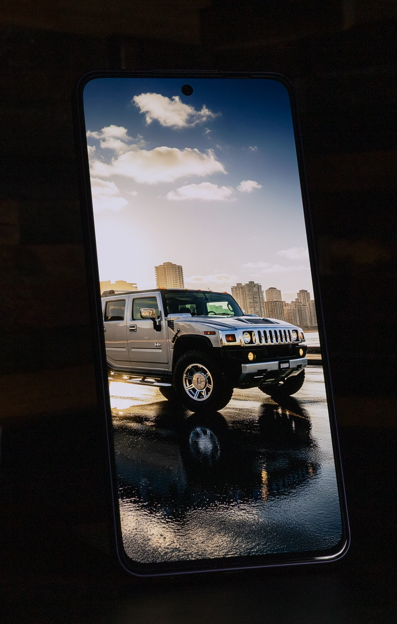 OPPO Reno13 Aのディスプレイに映し出された、水面に反射するSUVと都会の風景画像