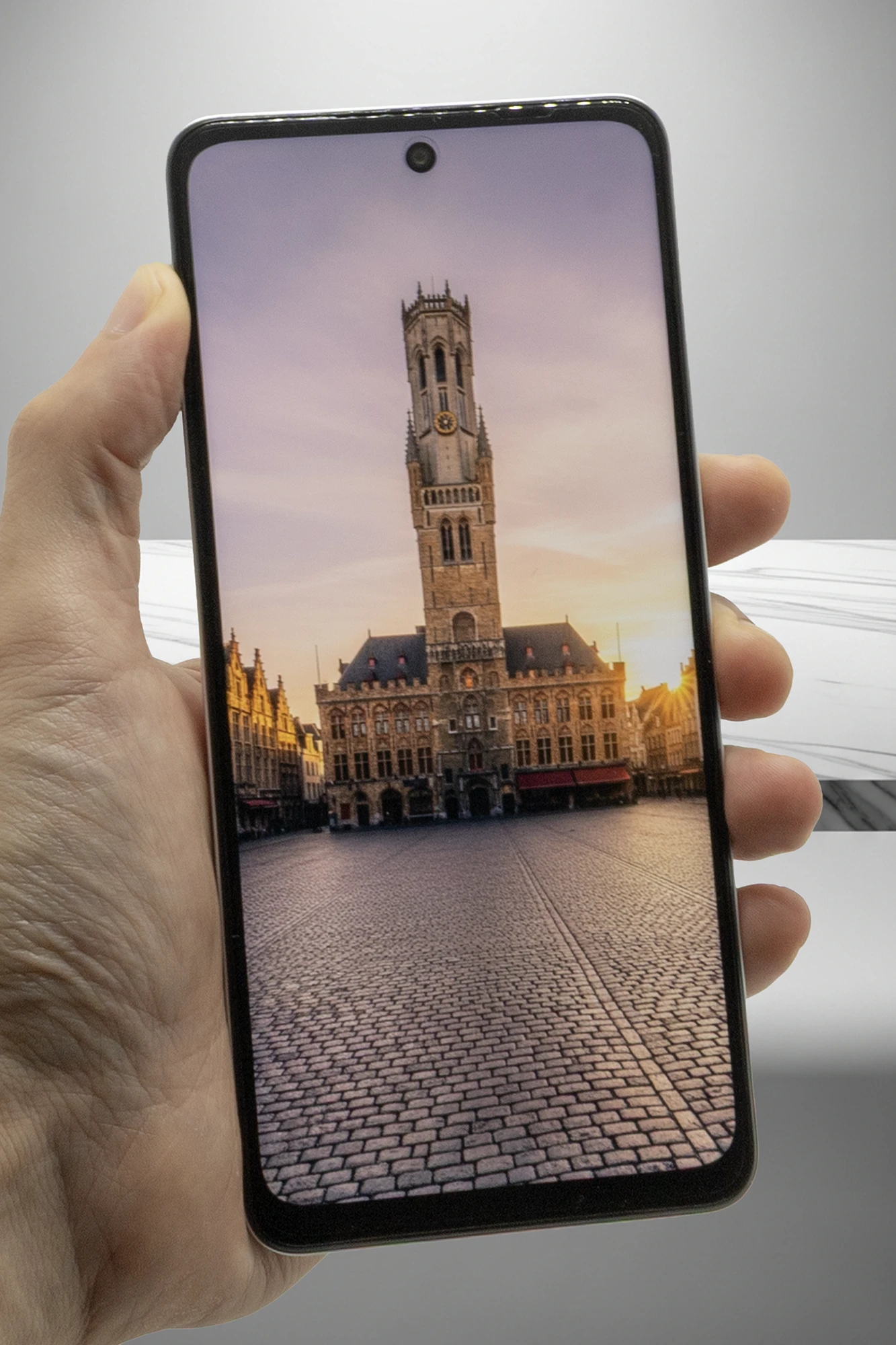 OPPO A5 5Gのディスプレイに、ベルギーのブルージュにある鐘楼と石畳の広場の写真が表示されている。ディスプレイ上部中央にはパンチホール型のフロントカメラが見える。