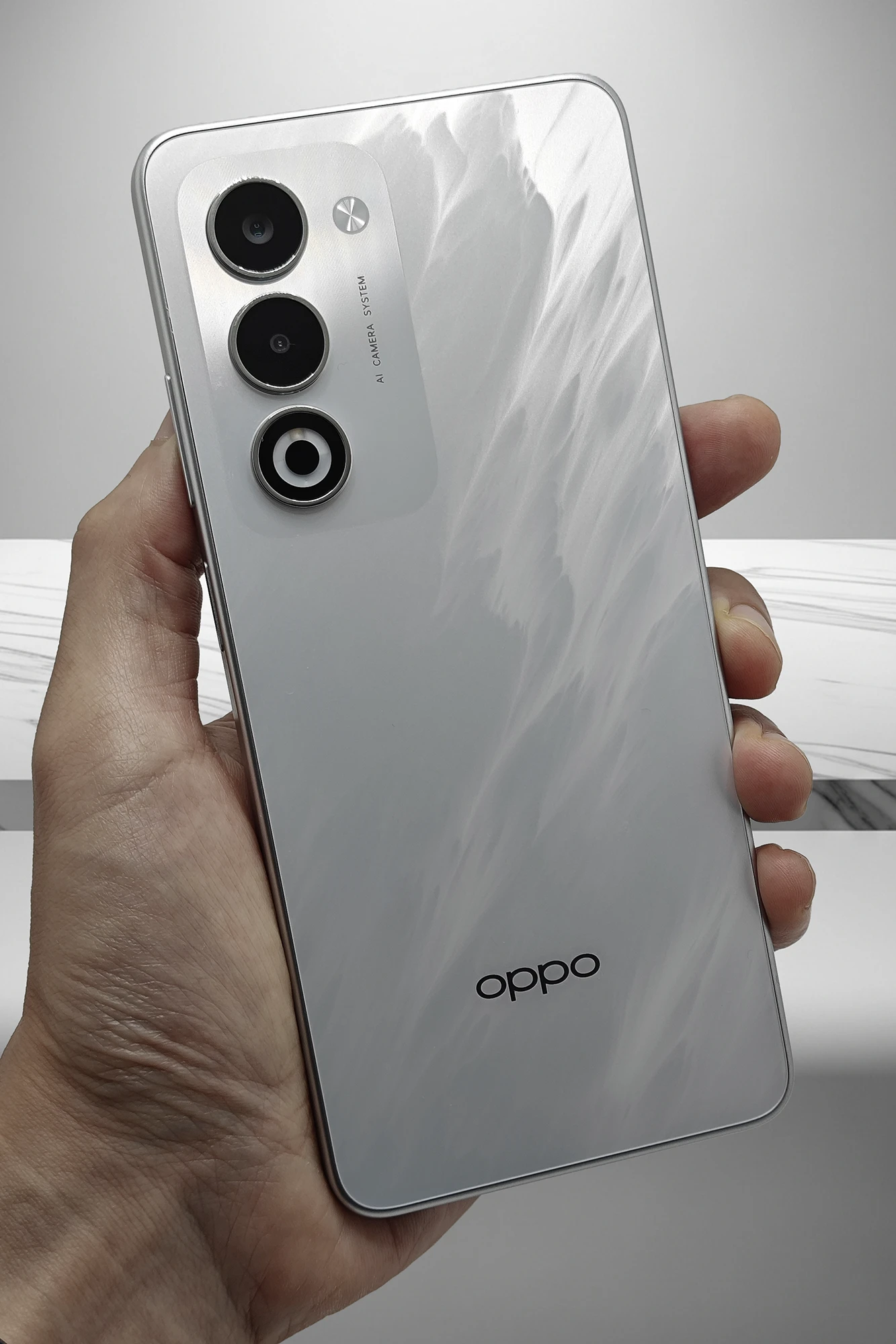 手に持たれたシルバーまたはホワイトのOPPO A5 5Gの背面。波状のテクスチャが特徴的で、縦に配置されたトリプルカメラユニットと「AI CAMERA SYSTEM」の文字、黒いOPPOロゴが見える。