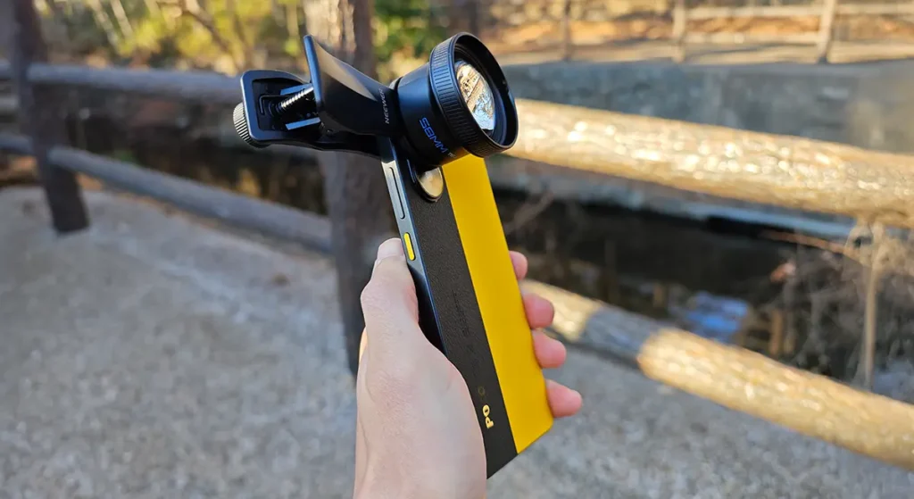公園の木製手すりを背景に、NEEWER 58mm望遠レンズを装着したPOCO X7 Proを手に持つ様子。屋外撮影でのサイズ感。