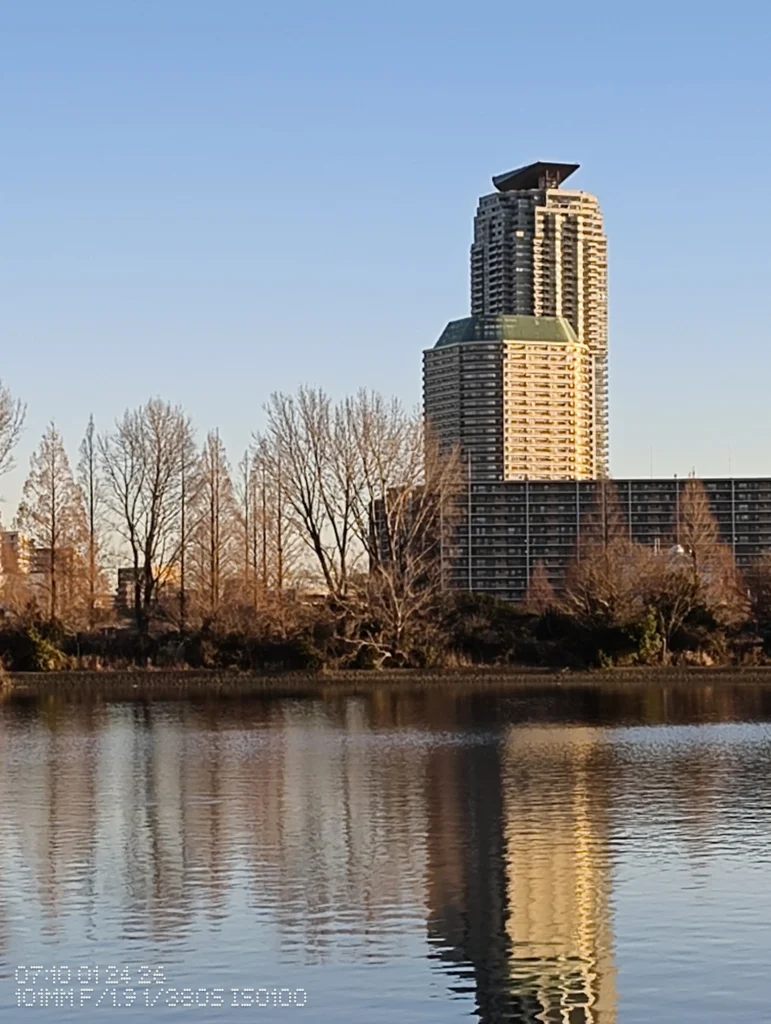 Nothing Phone (3a) Lite/カメラ:メイン(広角)約4.2倍ズーム/焦点距離:101mm相当/内容:高層ビルの階層構造と外周ラインを中心に据え、岸辺の木立と水面の揺らぎが圧縮感を強める