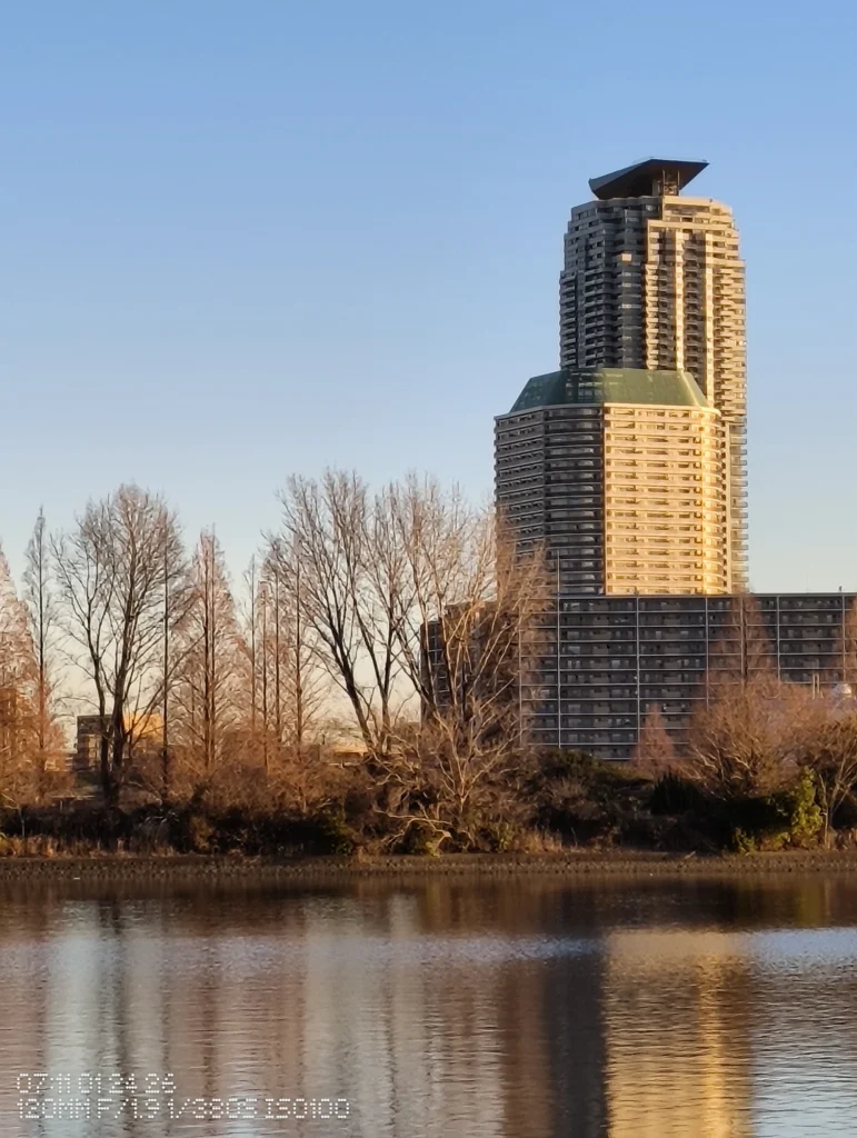 Nothing Phone (3a) Lite/カメラ:メイン(広角)5倍ズーム/焦点距離:120mm相当/内容:ビルの上層部とベランダの反復を大きく捉え、前景の木立と湖面の反射が背景を整理して距離感を出す