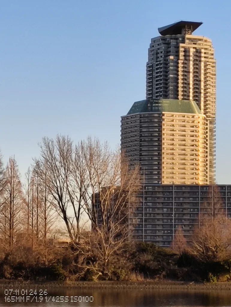 Nothing Phone (3a) Lite/カメラ:メイン(広角)約6.9倍ズーム/焦点距離:165mm相当/内容:窓列と外壁の面を中心に切り取り、木立のシルエットと湖面の反射が細かな揺れとして残る高倍率ズーム