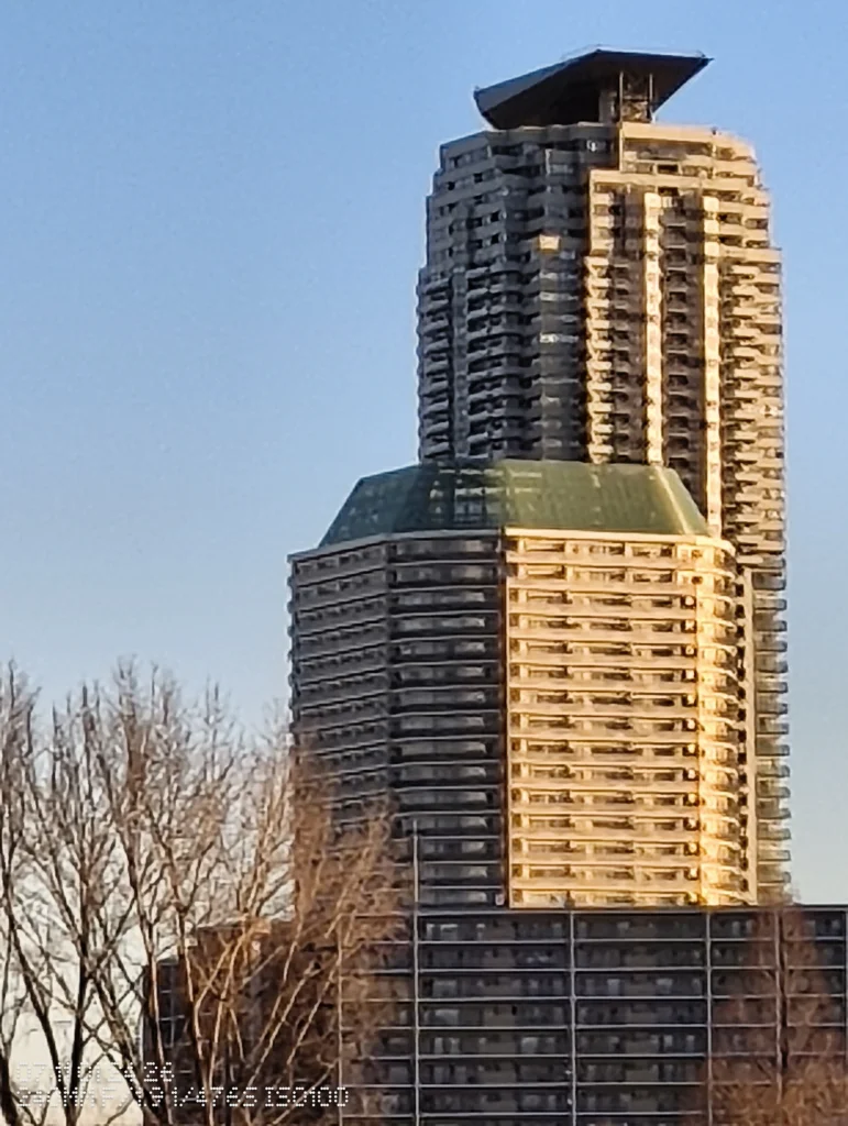 Nothing Phone (3a) Lite/カメラ:メイン(広角)10倍ズーム/焦点距離:240mm相当/内容:高層ビルの屋上付近まで大きく拡大して外壁の反復パターンを見せ、青空と木立が圧縮感を引き立てる