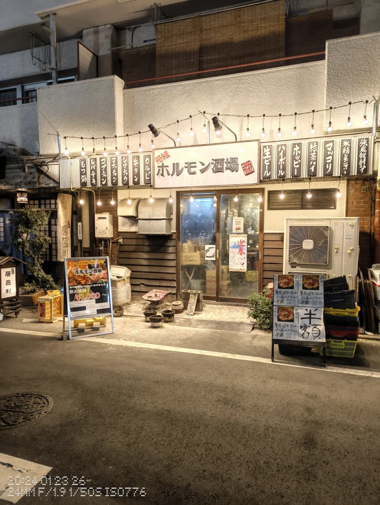 Nothing Phone (3a) Lite/カメラ:メイン(広角)/焦点距離:24mm相当/内容:電球列と看板が並ぶ店舗外観、入口のガラス扉とメニュー掲示が見える夜の飲食店街。