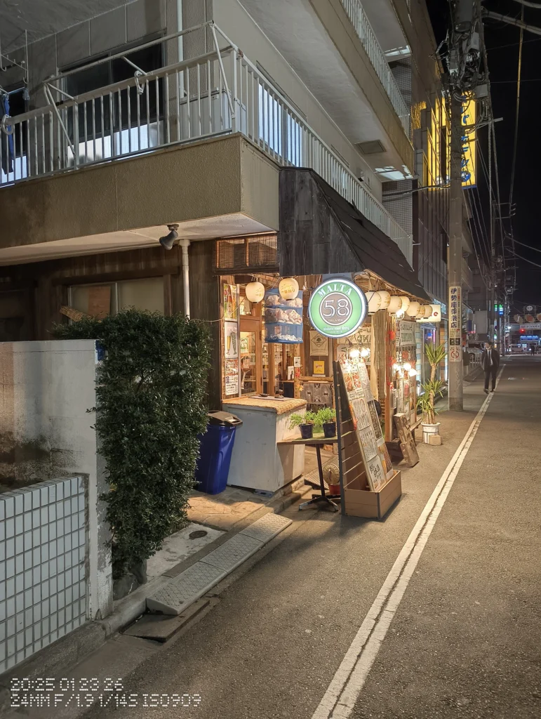 Nothing Phone (3a) Lite/カメラ:メイン(広角)/焦点距離:24mm相当/内容:電球列と看板が並ぶ店舗外観、入口のガラス扉とメニュー掲示が見える夜の飲食店街。