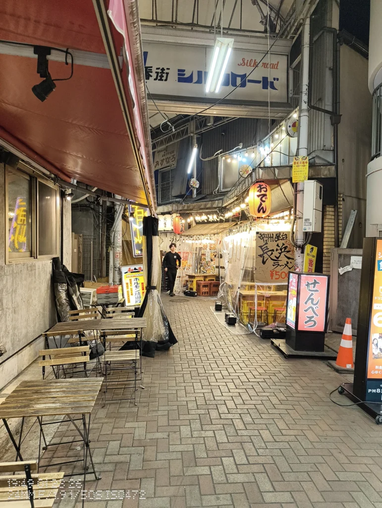 Nothing Phone (3a) Lite/カメラ:メイン(広角)/焦点距離:24mm相当/内容:アーケード内の提灯と蛍光灯に照らされた飲食店の並ぶ通路、テーブルや看板が続く夜景。