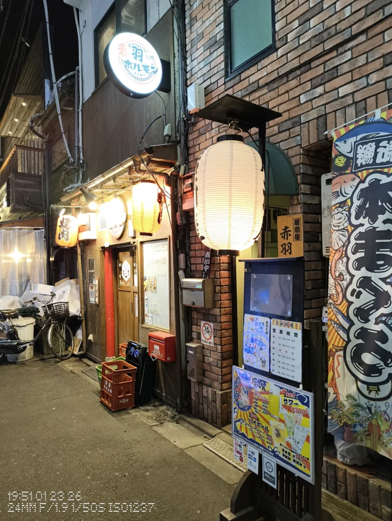 Nothing Phone (3a) Lite/カメラ:メイン(広角)/焦点距離:24mm相当/内容:レンガ壁の店舗入口に吊るされた提灯と看板、メニュー掲示や自転車が並ぶ夜の路地。