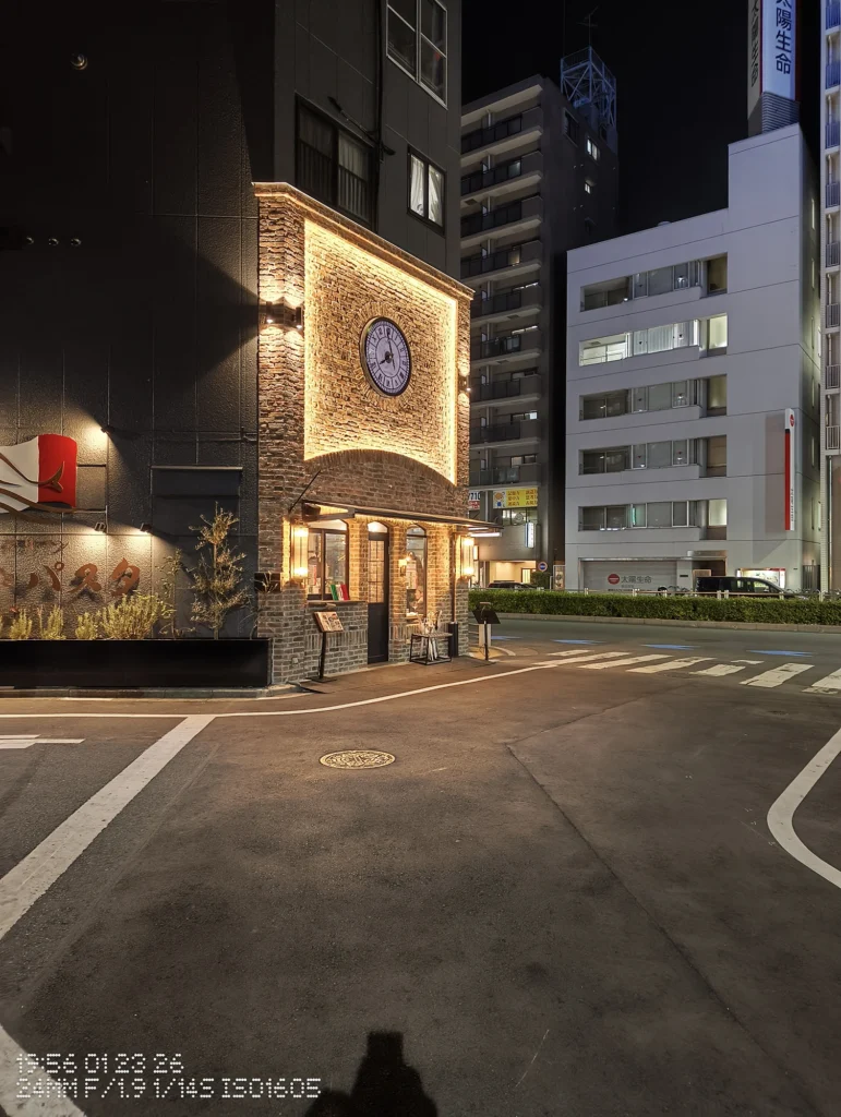 Nothing Phone (3a) Lite/カメラ:メイン(広角)/焦点距離:24mm相当/内容:交差点角のレンガ壁と大きな時計がライトアップされた夜景、横断歩道と周囲のビル明かりが見える。