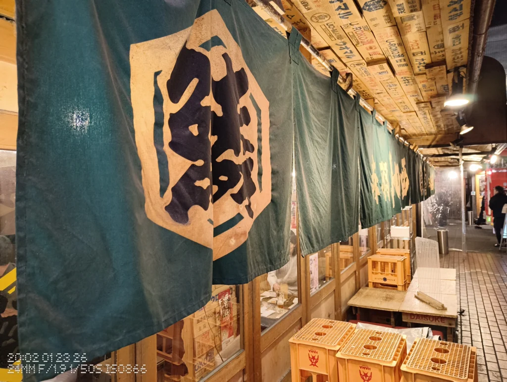 Nothing Phone (3a) Lite/カメラ:メイン(広角)/焦点距離:24mm相当/内容:店頭に掛かった大きなのれんと店内の暖色照明、通路に置かれた箱やテーブルが続く夜の飲食街。