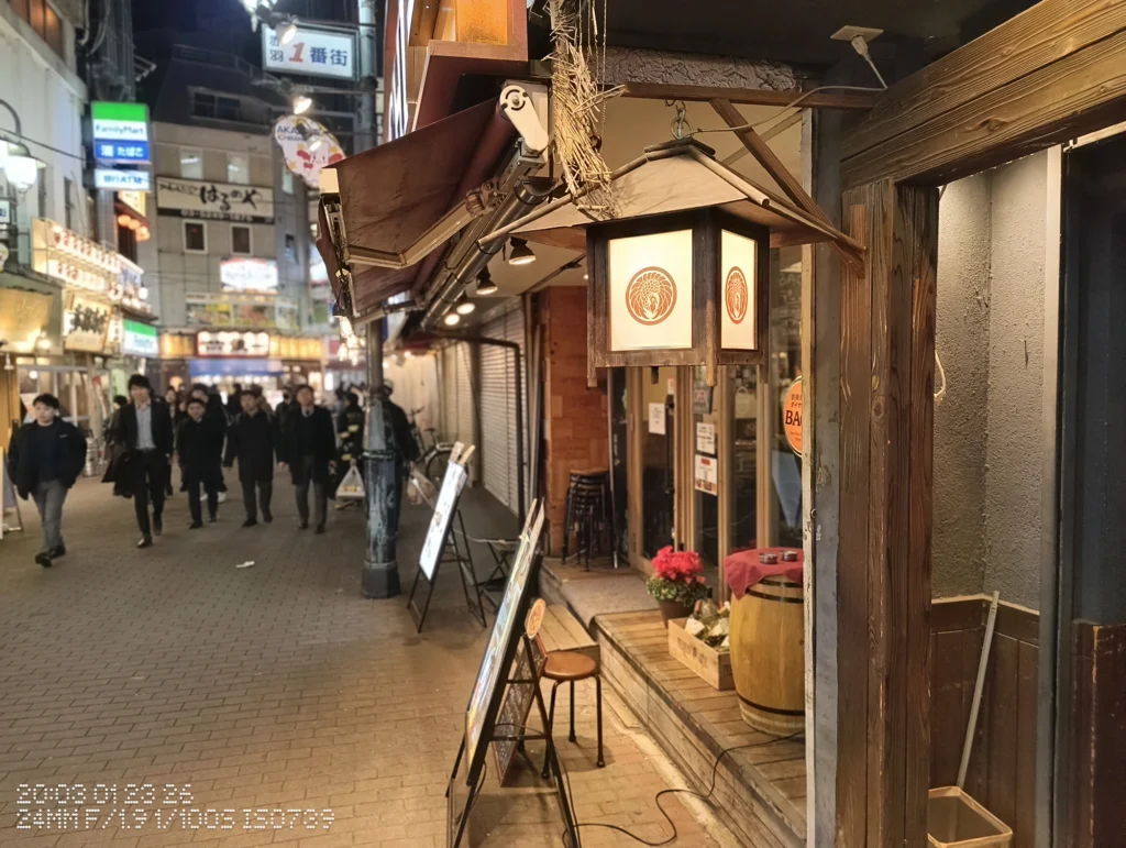 Nothing Phone (3a) Lite/カメラ:メイン(広角)/焦点距離:24mm相当/内容:木枠の行灯が店先で灯る夜の通り、入口周りの掲示物と奥に歩行者が見える飲食店街。