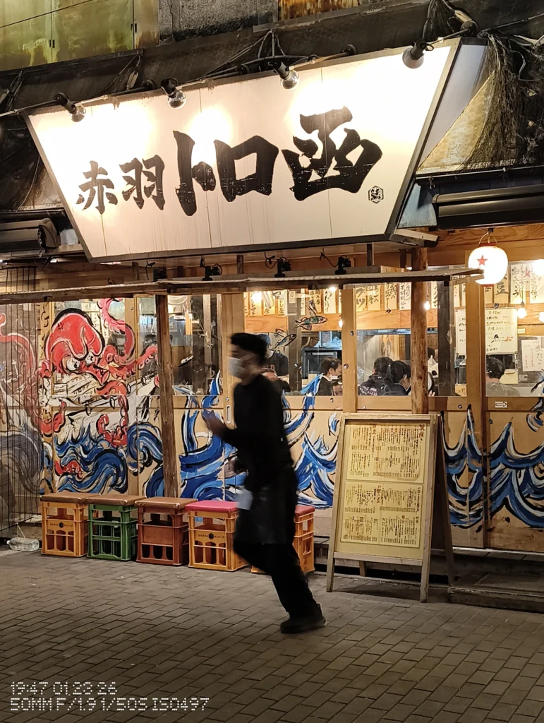 Nothing Phone (3a) Lite/カメラ:メイン(広角)2倍ズーム/焦点距離:50mm相当/内容:夜の路地で飲食店の発光サインと提灯が目立ち、店先を横切る人物の動きも写る光源多めの街並み。