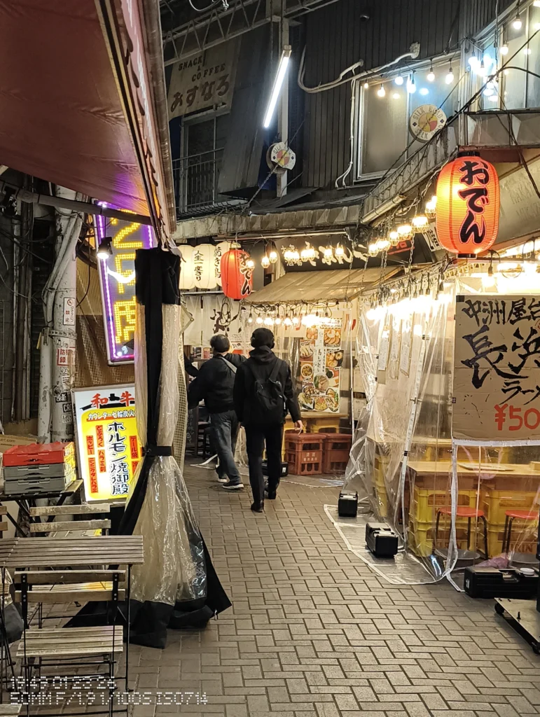 Nothing Phone (3a) Lite/カメラ:メイン(広角)2倍ズーム/焦点距離:50mm相当/内容:夜の飲食街で提灯とネオンが連なり、歩く人影を奥へ導く路地の光景を2倍ズームで引き寄せたスナップ。